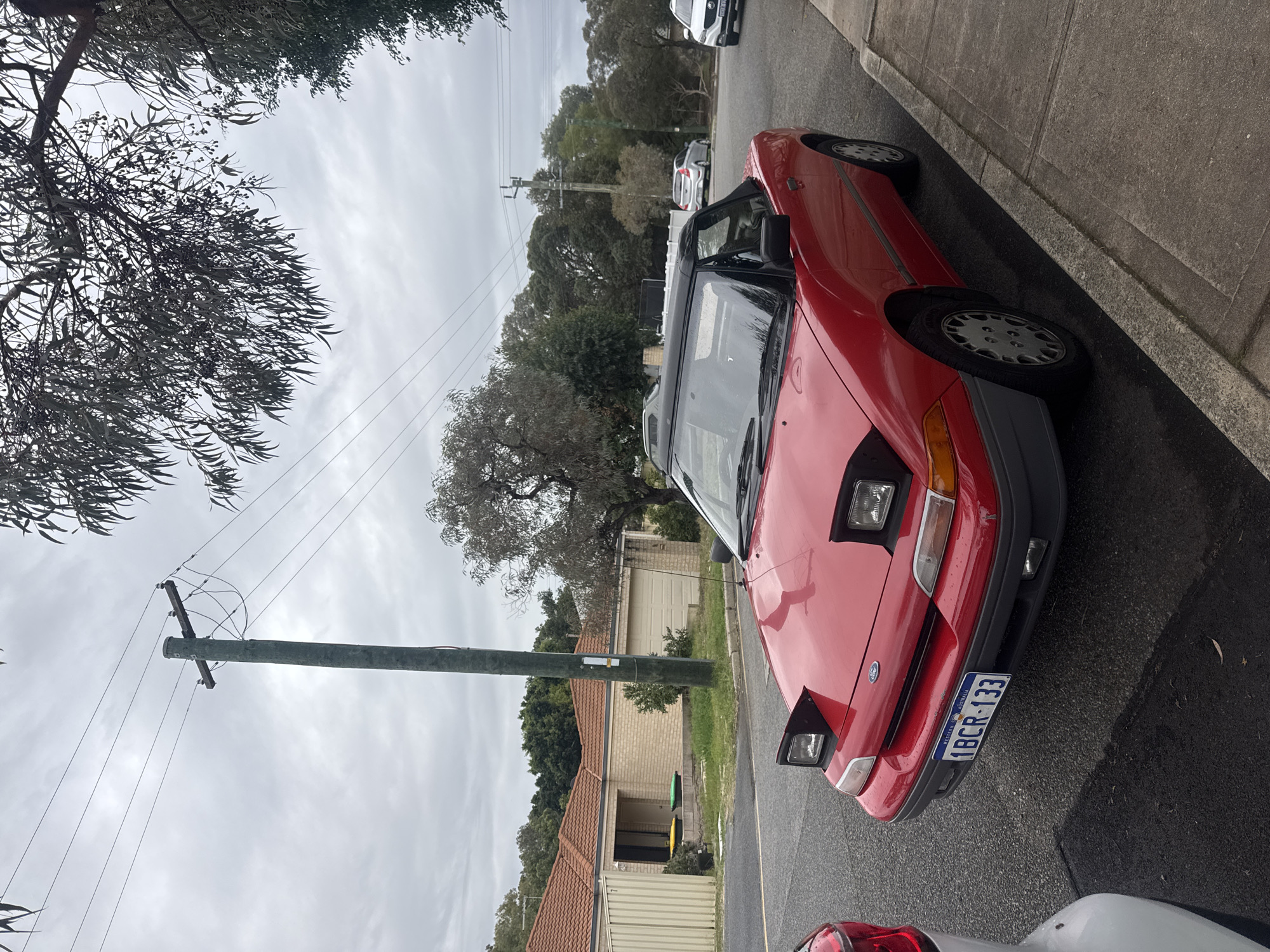 The image shows a red car parked on a street in a residential area. The license plate reads "1BCR-133," which indicates that the vehicle is registered in Western Australia. The car is parked next to a curb with a telephone pole nearby. The sky is overcast, and the trees suggest that the scene is located in a suburb. Based on the license plate, the location is likely within the state of Western Australia, but without additional information or visual cues specific to the area, it is difficult to conclusively pinpoint the scene as Bassendean.