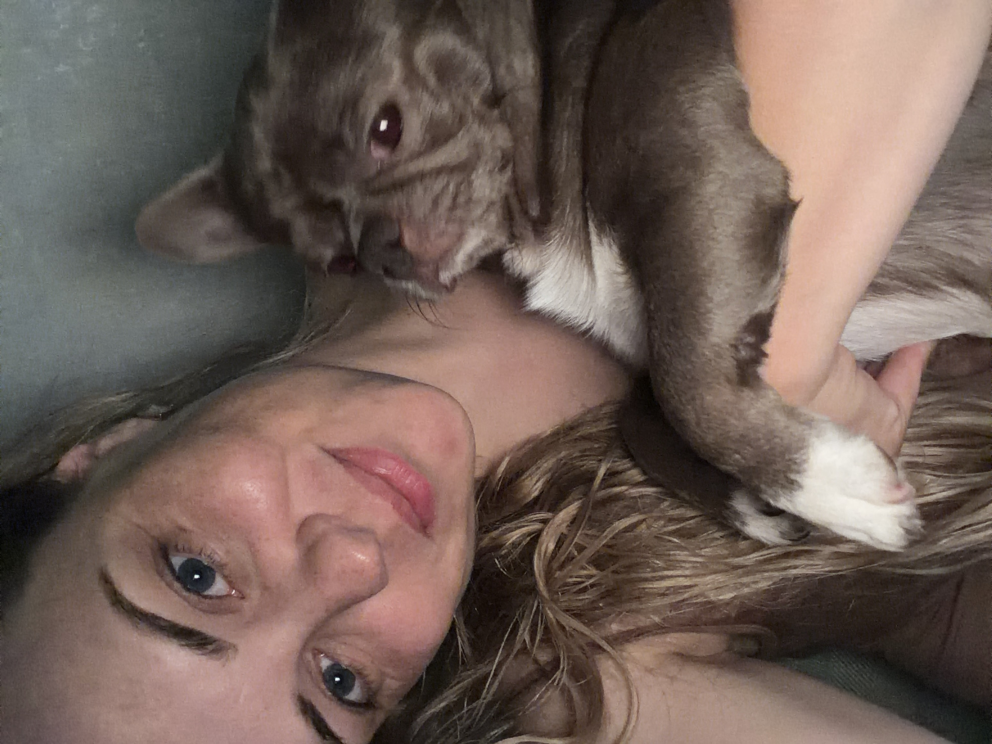A woman with light complexion, blonde wavy hair, and blue eyes is lying down and holding a brown and white bulldog-type dog. The dog's face is close to the woman's neck. The woman's face is turned slightly to the right, and she is looking directly at the camera with a slight smile. The background is a plain gray-green color. It is unclear if this photo was taken in Votkinsk, Russia.