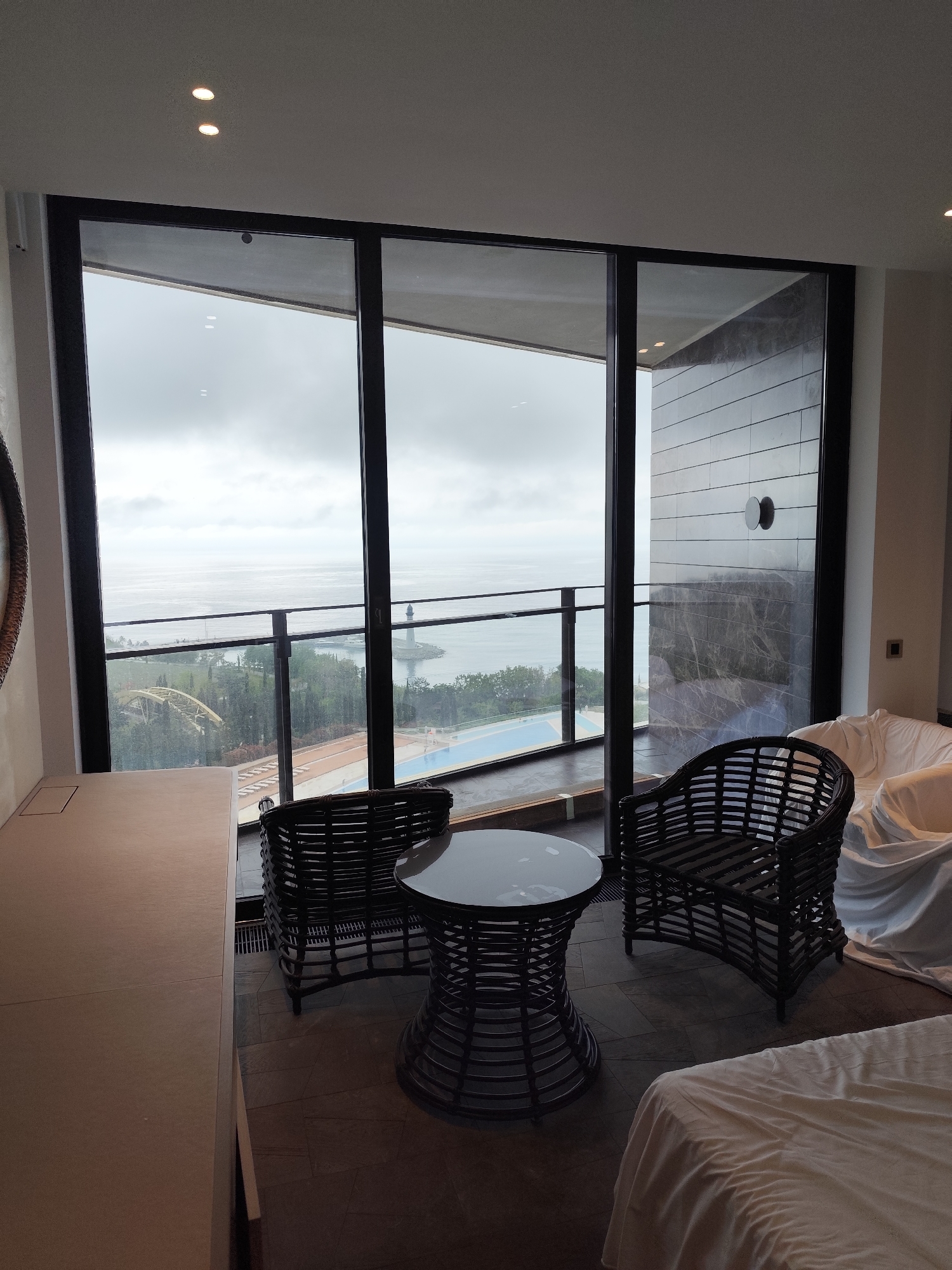 The image shows the interior of a room with a view of the sea. The room has a table and two wicker chairs. There is a large window with a view of the sea, a bridge, and a lighthouse. The sky is cloudy. The room appears to be located in the city of Unknown, Unknown.