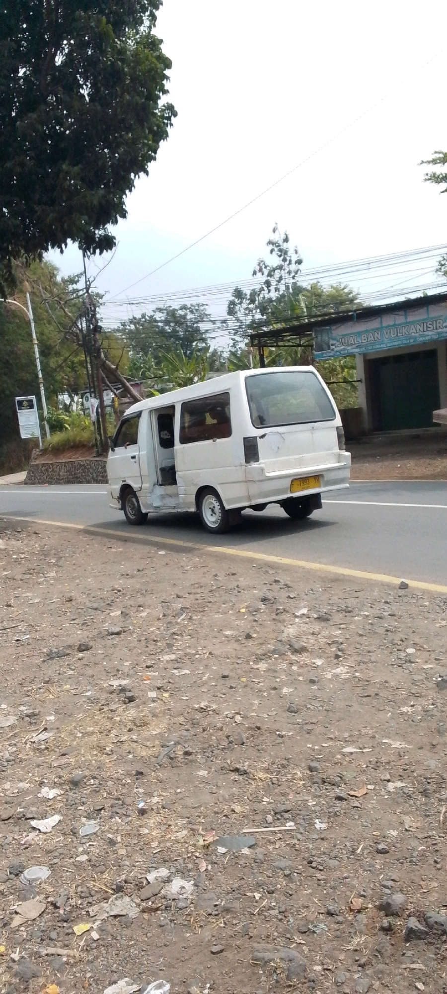 An outdoor, daytime, eye-level, medium shot captures a white van with its passenger door ajar, parked on the side of a paved road in Cilaku, Indonesia. The van, a Mitsubishi L300, exhibits signs of wear and tear, with rust visible along the lower body panels. Its license plate, a yellow Indonesian plate with the characters "F 1993 TH," is clearly visible on the rear bumper.
The road is bordered by a dirt shoulder littered with small rocks, dried vegetation, and debris, including what appears to be discarded plastic. To the left of the van, a steep, vegetated embankment rises, topped by a streetlamp and a signpost with text that is difficult to decipher due to image quality. Telephone poles and tangled wires are prominent features of the background, suggesting an urban or semi-urban environment.
Across the road, a low building with a corrugated metal roof and a dark green garage door stands. A sign above the building reads "JUAL BAN VULKANISIR," indicating a tire repair shop. Lush green foliage and trees are scattered throughout the scene, indicating a tropical climate. The sky is overcast, with a hazy white appearance. No people are visible in the frame. The overall scene depicts a common roadside view in a developed area of Indonesia.