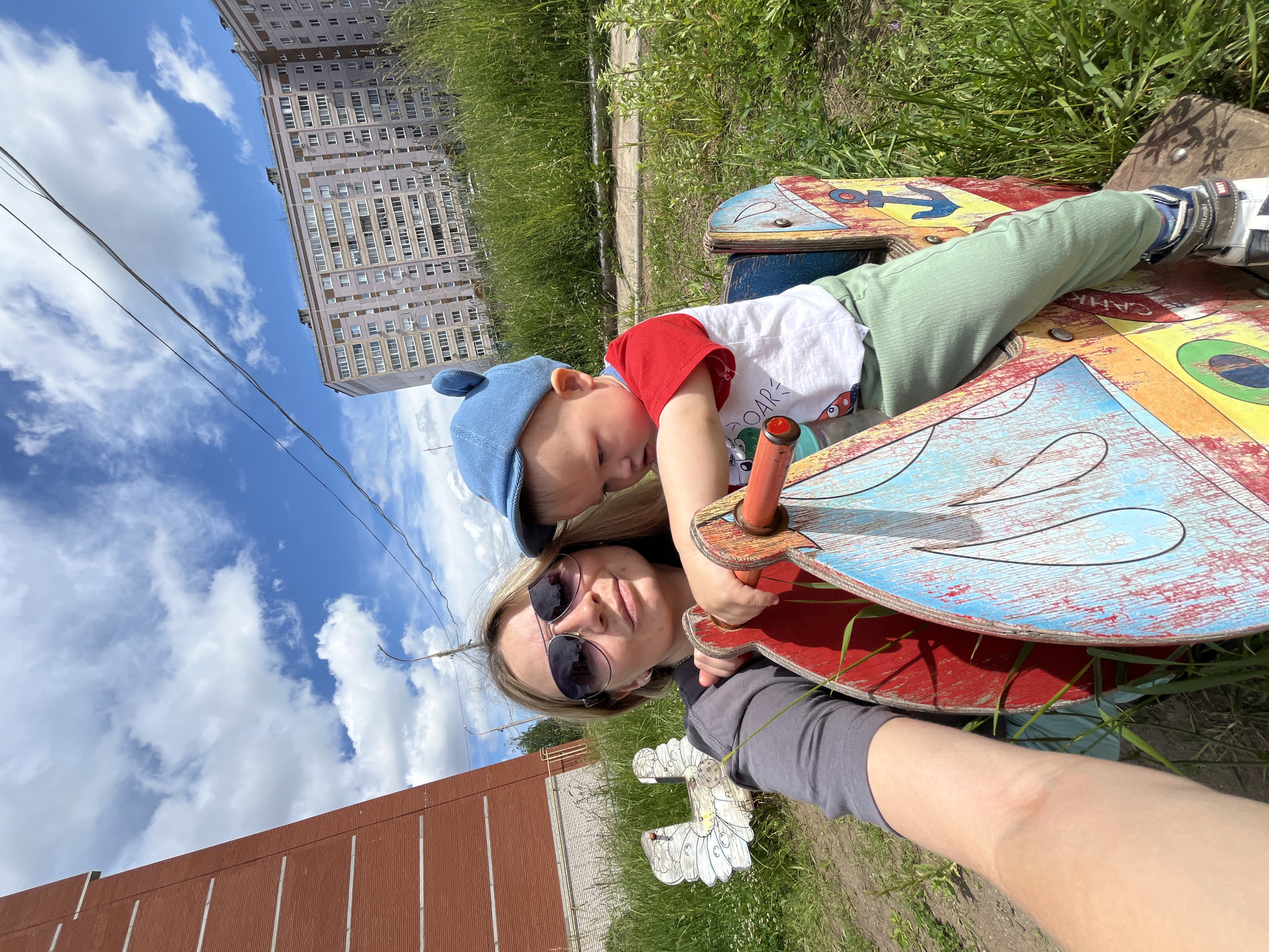 In the city of Chaikovsky, Russia, a woman wearing sunglasses and a grey long-sleeved shirt poses for a selfie with a young child in a blue cap and a red and white t-shirt. The child is seated on a colorful wooden rocking toy. The background features a tall brick building on the left and a large apartment complex on the right, with tall green grass and a cloudy blue sky. A white wooden chicken-shaped ornament is visible to the left of the woman.