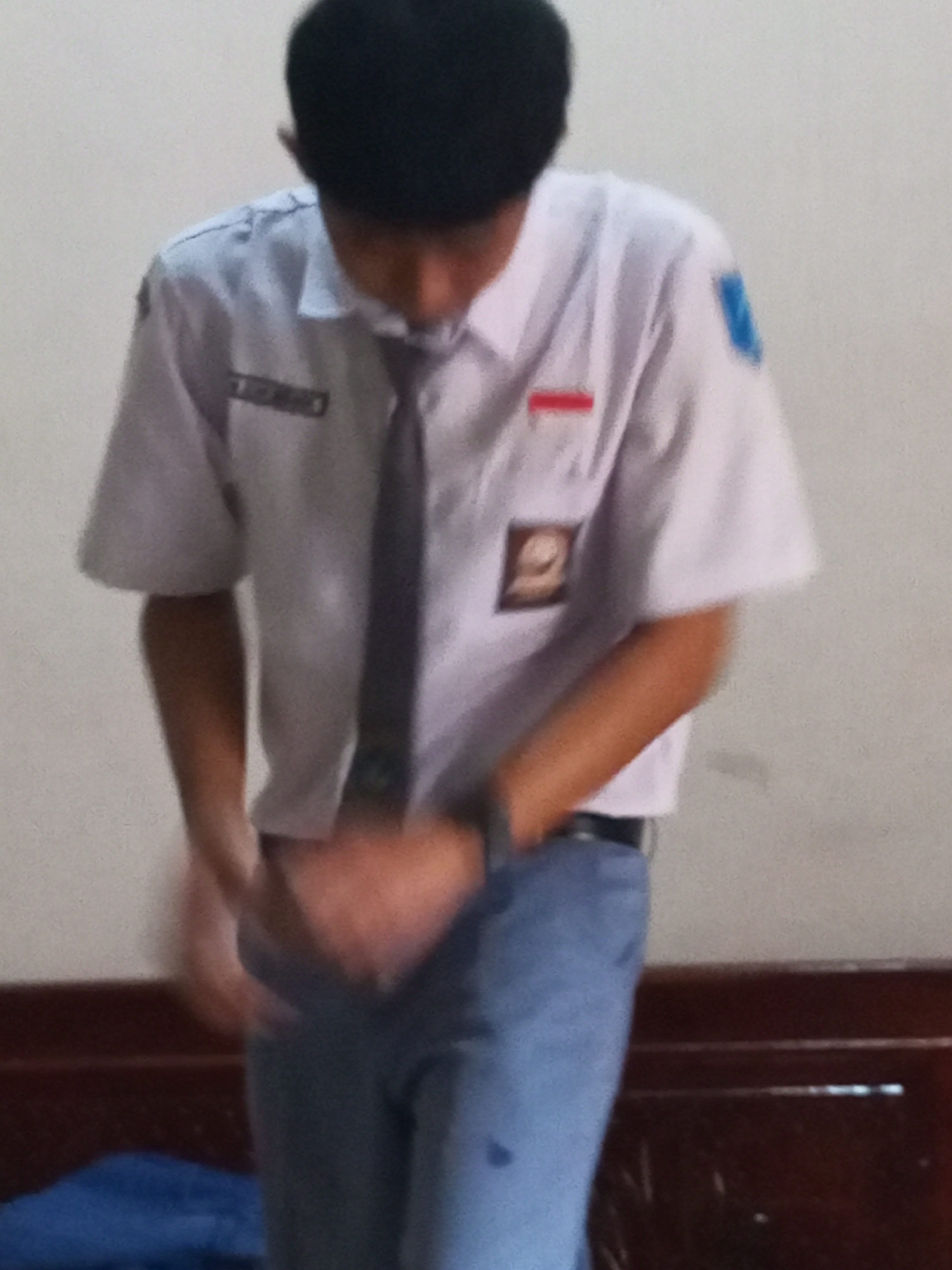 A young person wearing a school uniform, consisting of a white shirt with a tie and blue pants, stands facing downwards with their hands in front of their body. They have short black hair. The background is a plain, light-colored wall. The scene appears to be indoors.