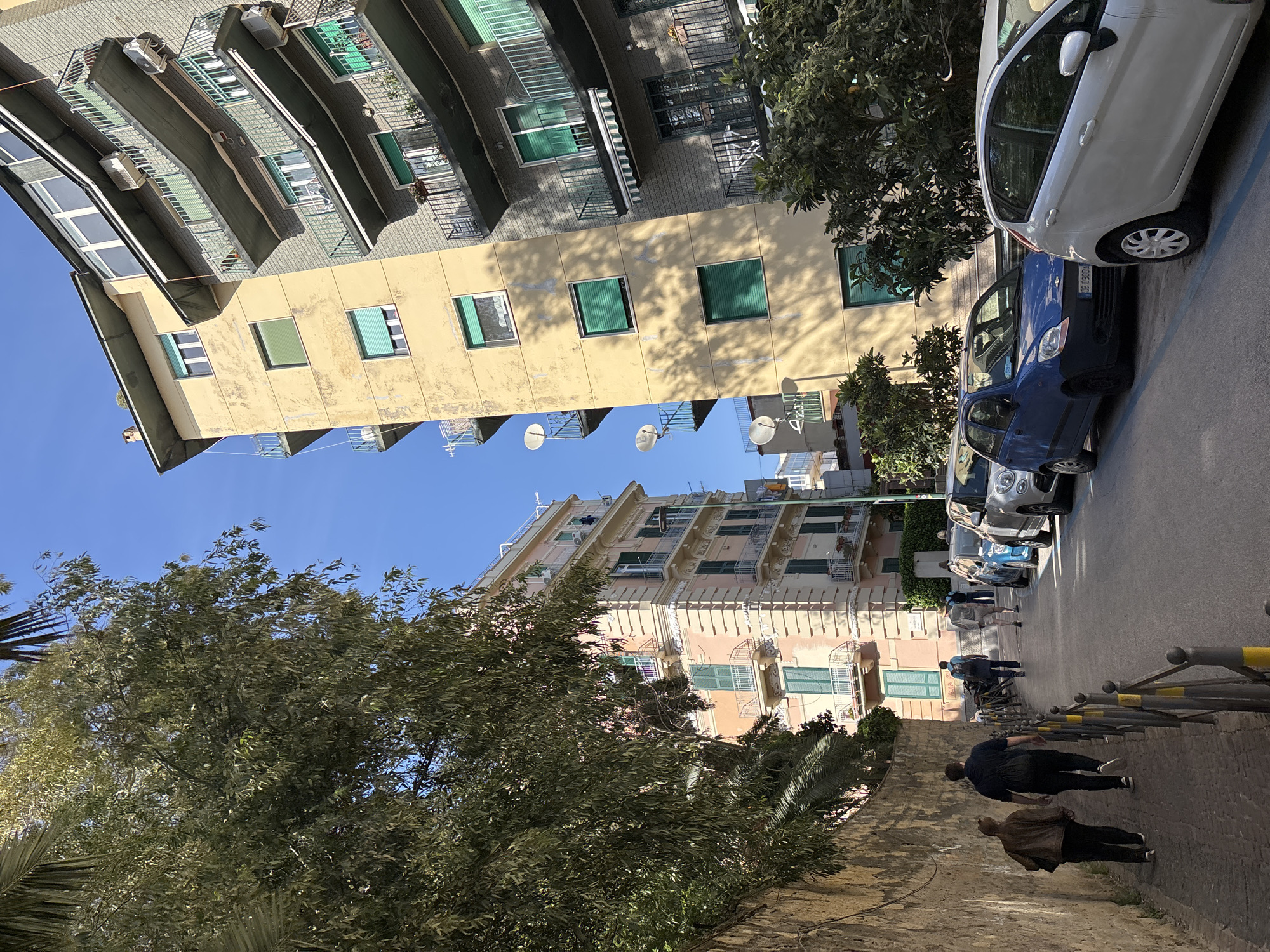 The image shows a street scene in an urban area, with buildings lining both sides of the street. On the right side of the street, several cars are parked, including a white car and a blue car. Pedestrians are walking along the sidewalk, some of them near a railing. The buildings vary in color, with one being yellow and another being pink. The sky is blue and clear. Trees and other vegetation are present in the foreground and throughout the scene. Satellite dishes are visible on the roofs of the buildings.