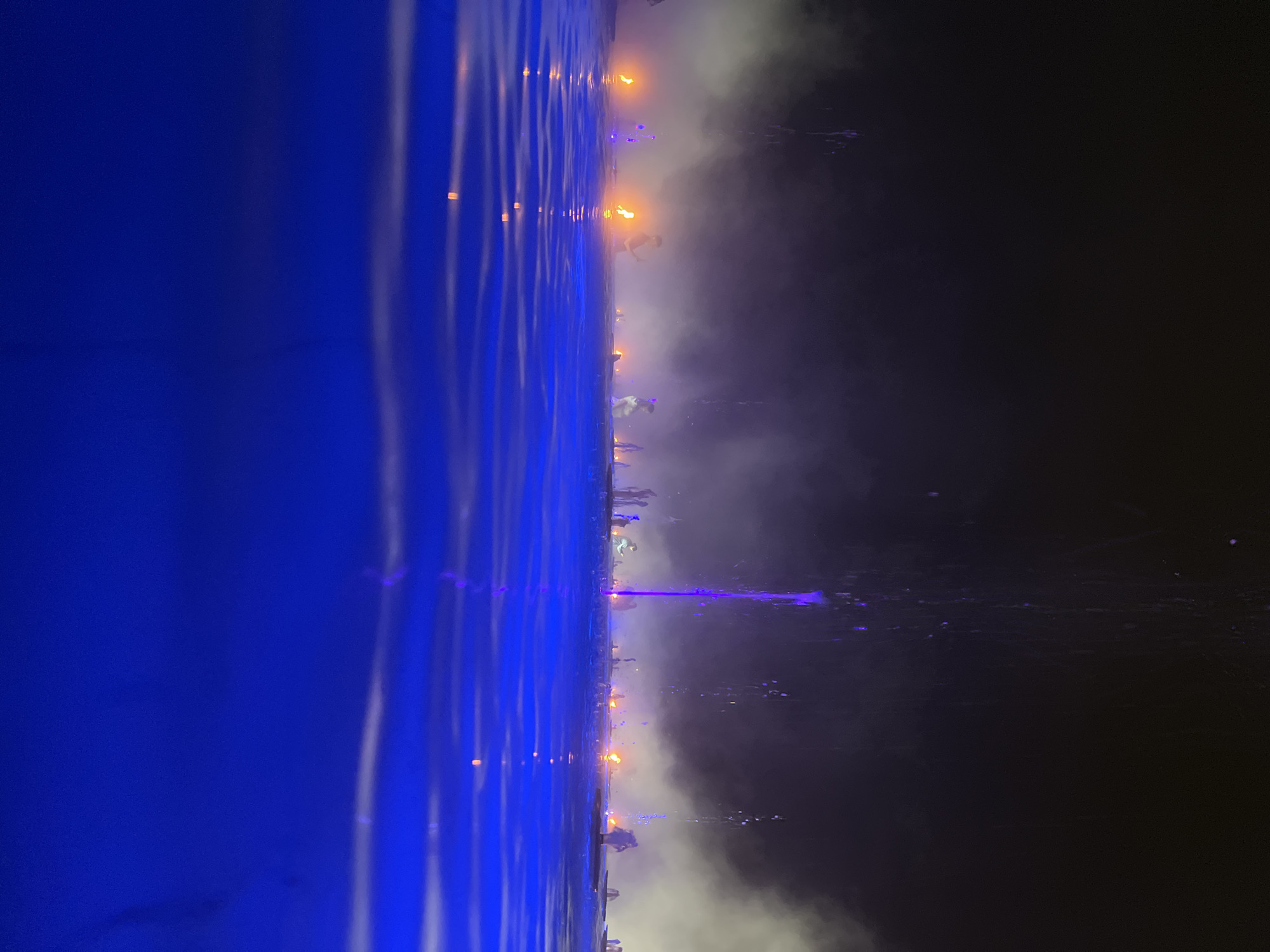 The image depicts a night scene at a water attraction, likely a fountain show, in Krasnodar, Russia. The foreground is dominated by deep blue water with visible ripples and reflections of lights. In the background, a dark sky serves as a backdrop for a series of illuminated elements. These include plumes of water from fountains, some of which appear to be lit with purple light, and several bright orange flames from torches or similar devices. Silhouettes of people can be seen standing amidst the display, some appearing to interact with the water or the lights. Wisps of white smoke or mist drift through the air, adding to the atmospheric effect of the show. The overall impression is of an outdoor entertainment event at night, characterized by water, fire, and light.