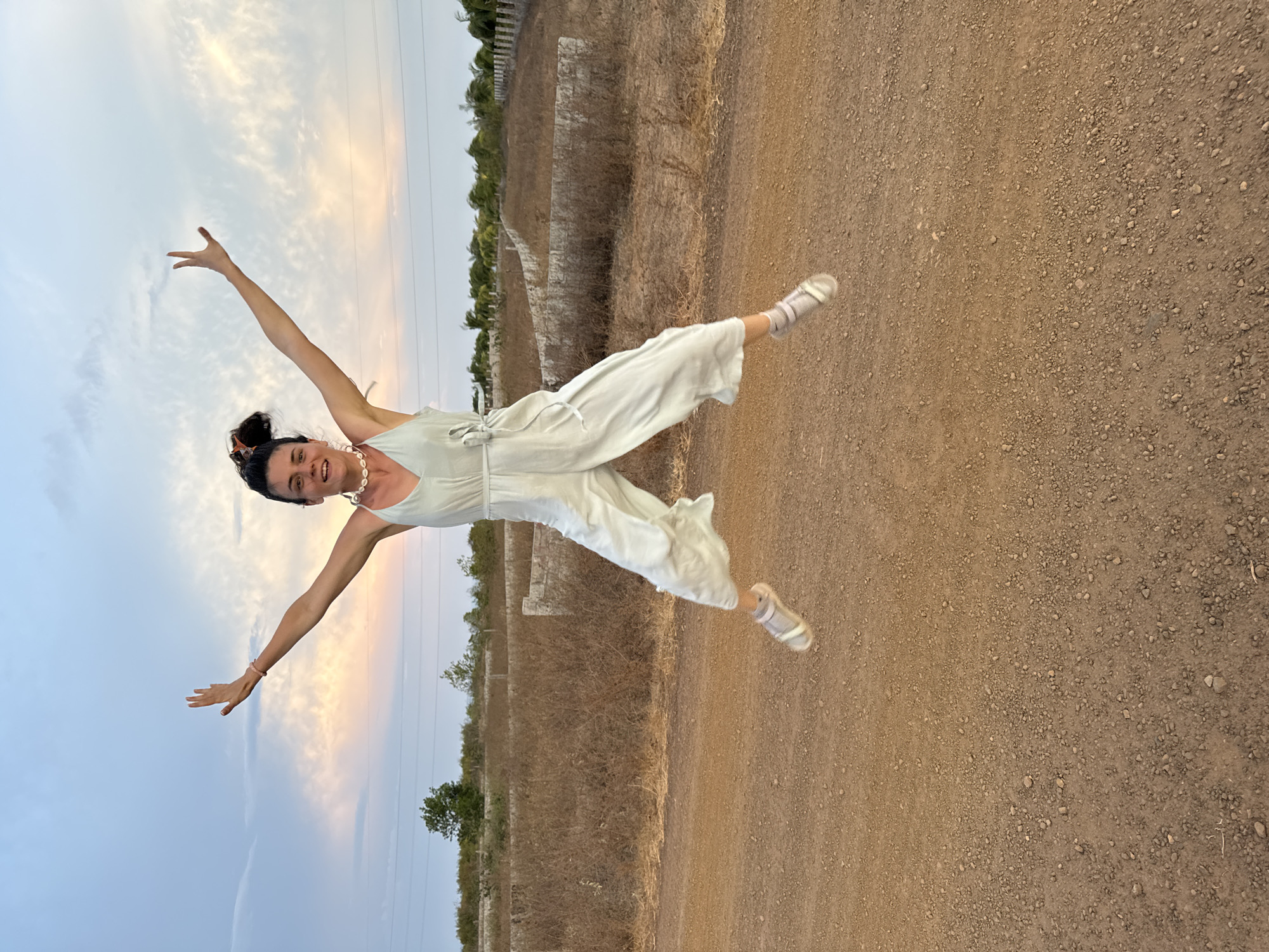 A woman is jumping in the air with outstretched arms and legs on a dirt road. She is wearing a light-colored jumpsuit, light-colored shoes, and a shell necklace. The dirt road is surrounded by dry grass and vegetation. In the background is a faded sunset sky. This image appears to be taken outdoors, although no specific location in Mũi Né, Vietnam is identifiable.
