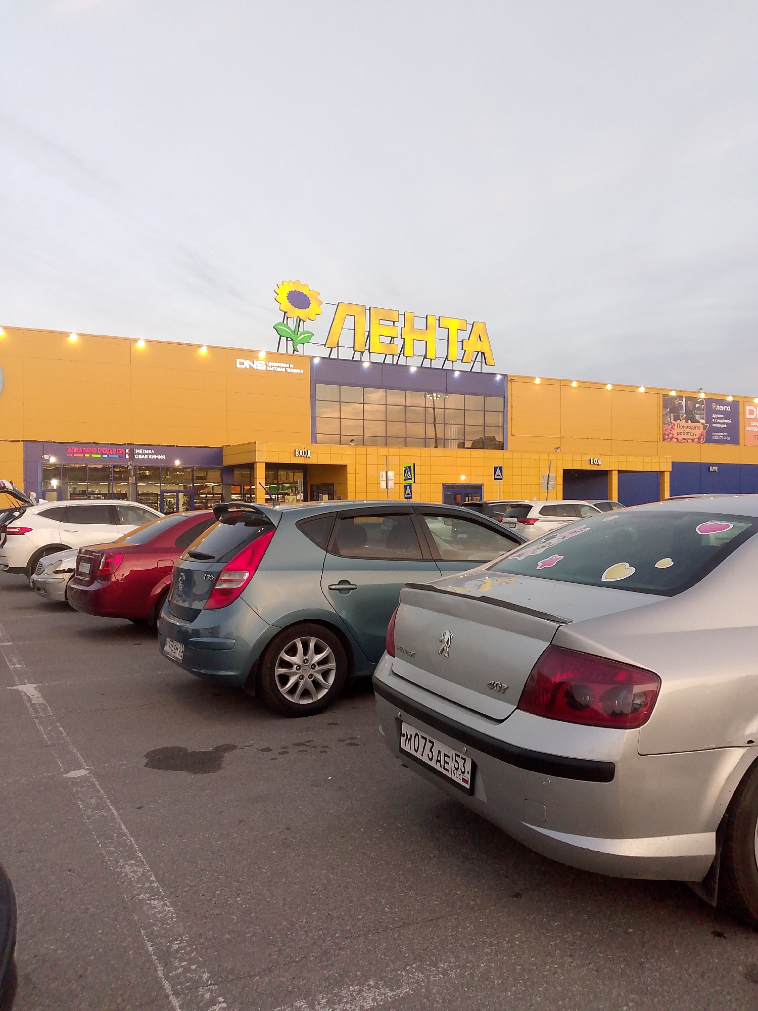 This is an outdoor, eye-level, medium shot of a parking lot in front of a large retail building. The building is painted yellow and has a large sign that reads "ЛЕНТА" in yellow lettering with a sunflower logo. The sky is overcast and a pale grey. Several cars are parked in the lot. In the foreground, a silver Peugeot 407 is parked, its rear bumper and taillights visible. To the left of the Peugeot, a teal Hyundai i30 is parked, its rear and side visible. Further to the left, a dark red sedan is parked, followed by a white SUV with its trunk open. The parking lot is paved with asphalt and marked with white lines. There are pedestrian crossing signs visible. The scene appears to be in Shimsk, Russia.