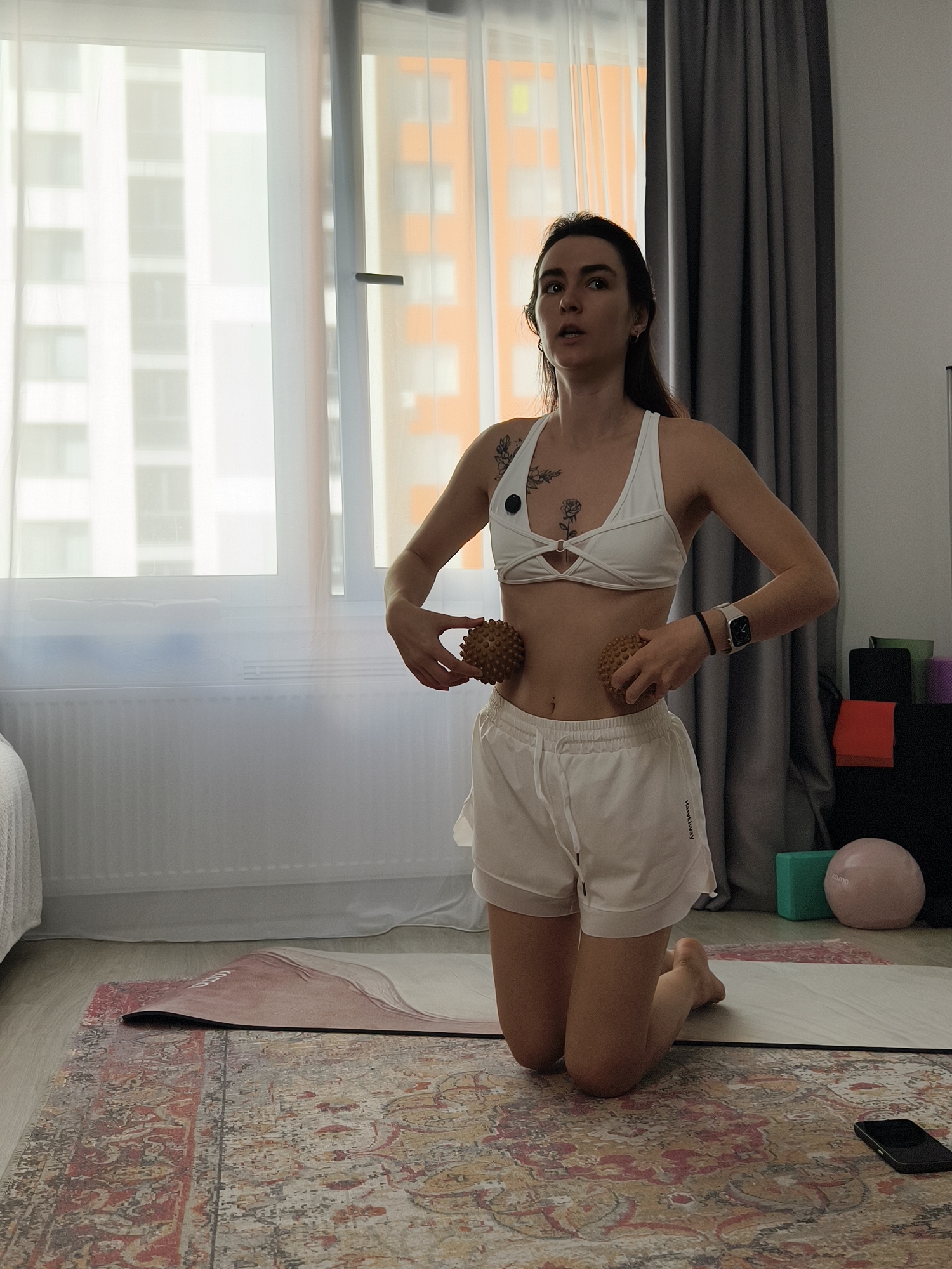 The image depicts a woman in white athletic wear, kneeling on a yoga mat and rug in what appears to be an indoor exercise or living space. She is holding two textured massage balls against her abdomen. She has long, dark hair and several tattoos on her chest. Behind her, a large window reveals a blurred view of apartment buildings, suggesting an urban setting. To the right, there are various fitness accessories, including yoga blocks and a pink ball. The overall scene suggests a personal fitness routine being performed at home. The presence of apartment buildings outside the window indicates a residential area within a city. While no specific landmarks are visible, the style of the buildings might be consistent with architecture found in cities like Tyumen, Russia, a regional capital known for its modern apartment complexes.