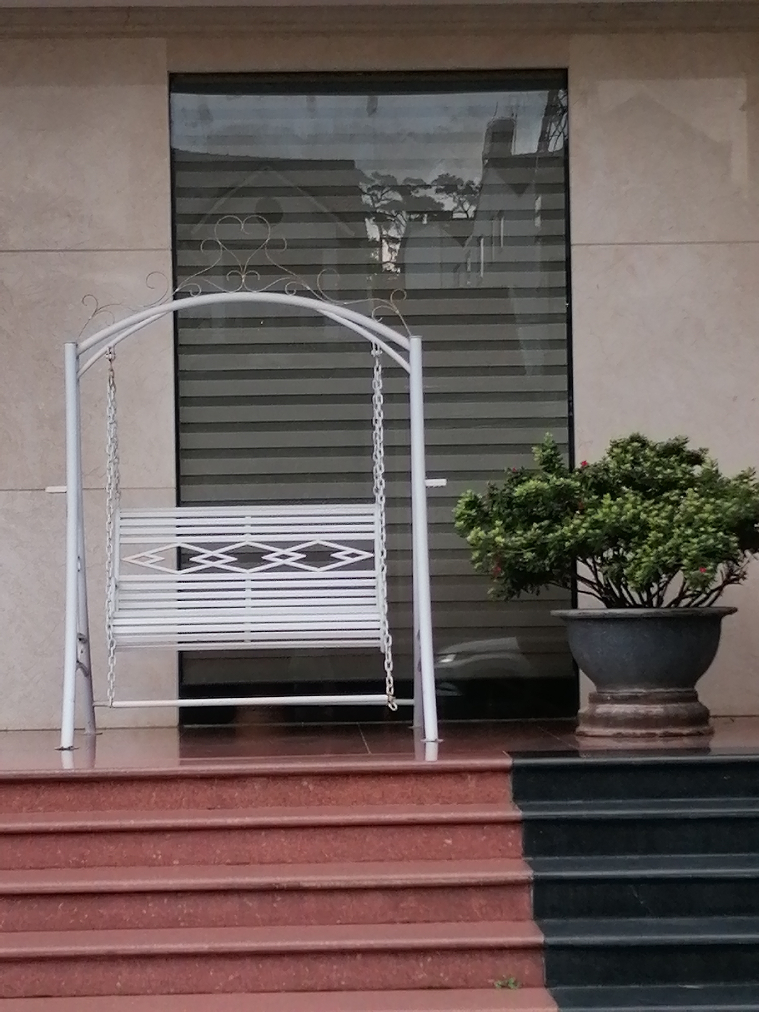 The image depicts a white metal swing set with a decorative arched top, positioned in front of a large window with horizontal blinds. The swing seat is white and features a geometric pattern. To the right of the swing, a potted green shrub is visible. The scene is set on a tiered staircase, with the lower steps being red and the upper steps being black. The building facade is a light beige color. A reflection of trees and structures can be seen in the window. The location is identified as Da Lat, Vietnam.