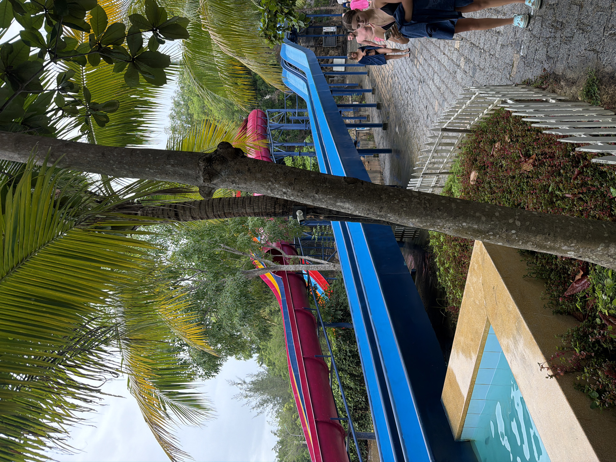 The image depicts a water park in Nha Trang, Vietnam, on a cloudy day. In the foreground, a bright blue water slide descends towards a small pool. To the left, palm trees and other lush green foliage frame the scene. In the background, a red and yellow water slide is visible, with a person in a dark swimsuit and towel in the mid-right of the frame, looking at their phone. Another person in a dark blue towel walks away from the camera, while a couple is seen walking away from the slides. The ground is paved with cobblestones and appears wet.