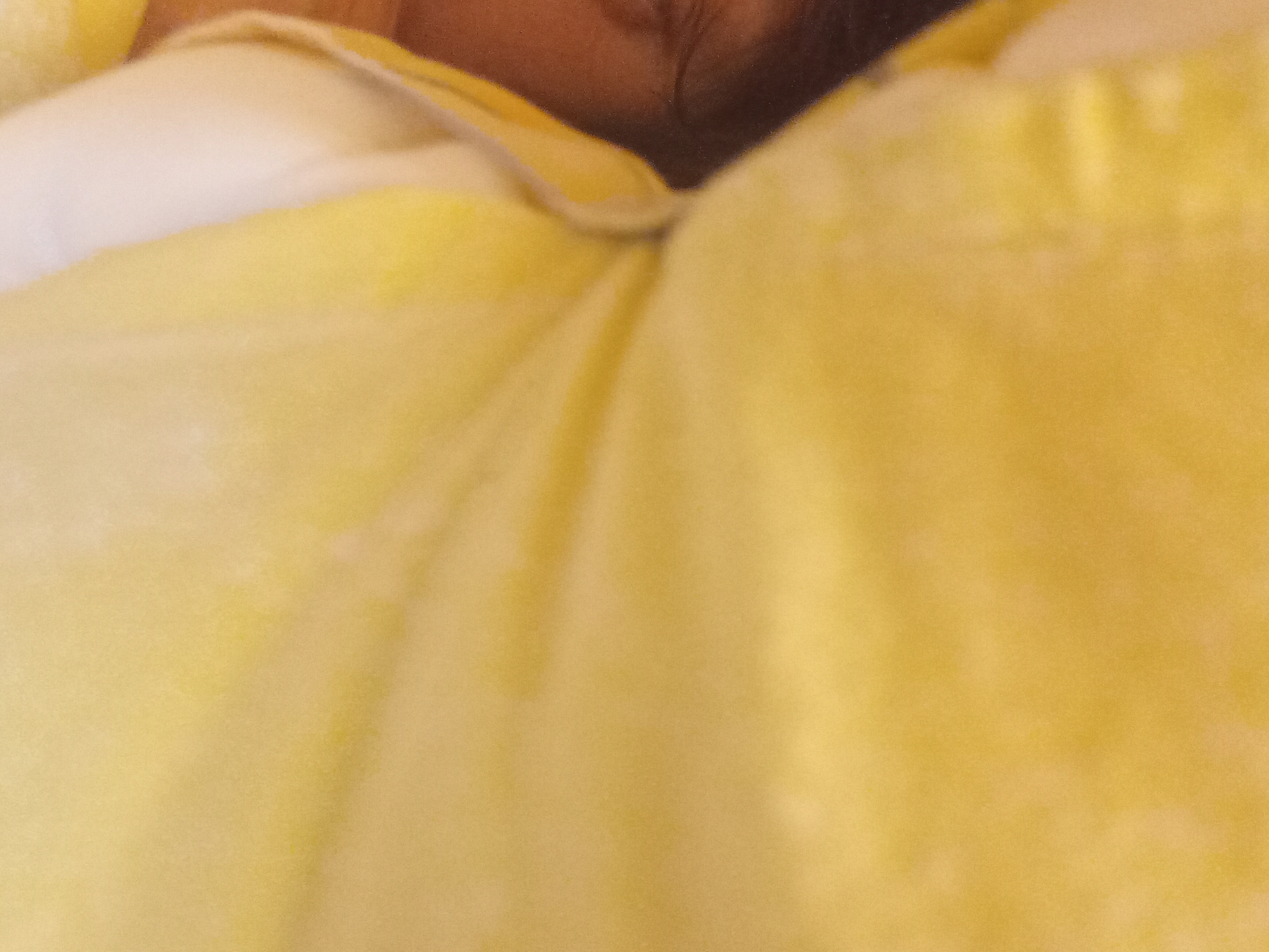 A close-up, blurry image shows a person's face, primarily their left eye and dark hair, against a bright yellow background. The yellow appears to be fabric, possibly a blanket or clothing, with some creasing and subtle variations in tone. The image is predominantly yellow with a small section on the left showing darker skin tones and a portion of an eye. The lighting is soft and diffused. There are no discernible environmental details or location indicators within Zuberec, Slovakia.