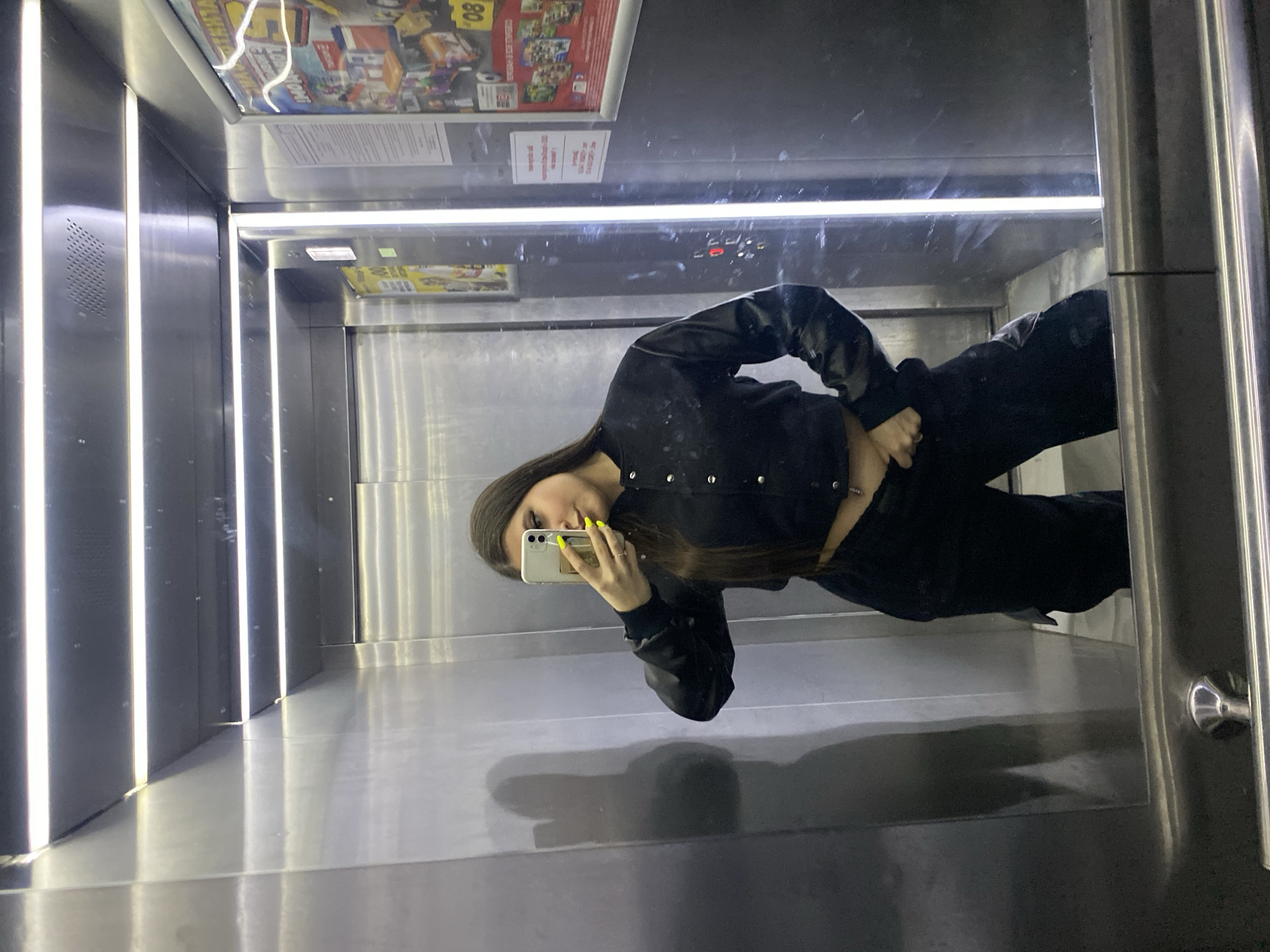 A young woman is seen in the mirror of an elevator, presumably in Chaikovsky, Russia, as indicated by the advertisement poster at the top. She is holding a white smartphone in her left hand, seemingly taking a selfie, and has her right hand on her hip. She is wearing a black jacket and black pants. The elevator is metallic and has vertical light fixtures.