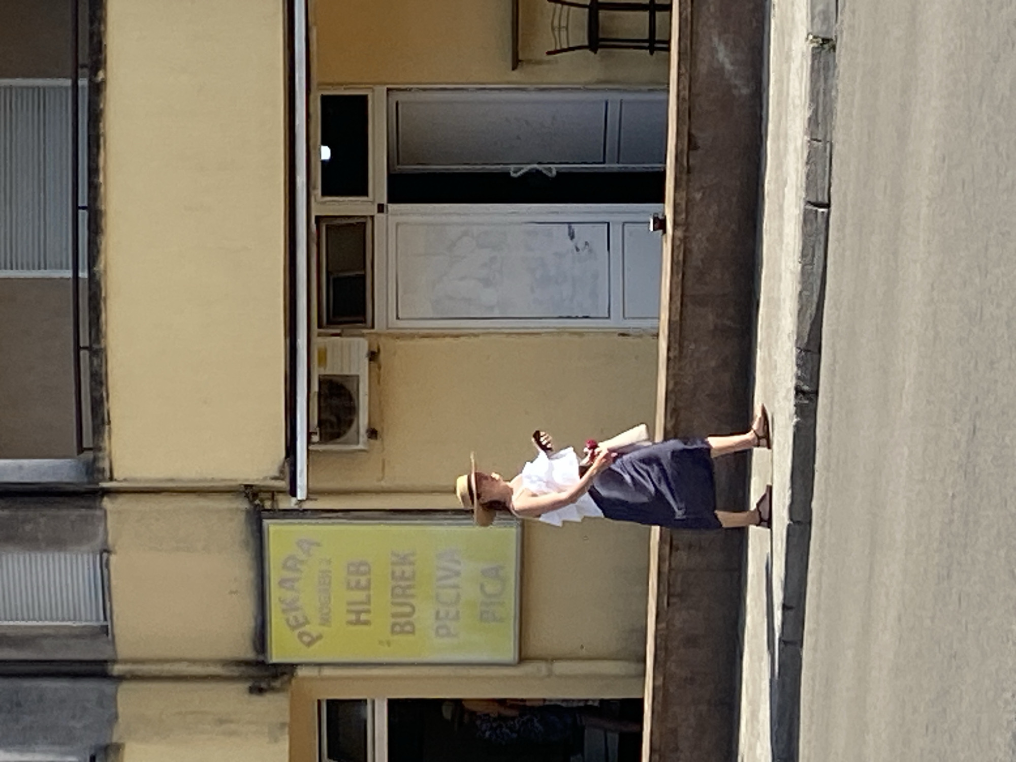 The image depicts a street scene in Budva, Montenegro, featuring a woman walking on a sidewalk in front of a yellow building with a sign that reads "PEKARA" (bakery). The sign also lists various baked goods and pizzas. The woman, wearing a white top and a dark skirt, is carrying a bag and a phone. She is wearing a straw hat and sandals. To the right of the entrance, there is an open door and a chair. A small cup is visible on a ledge in front of the building. The scene is illuminated by sunlight, casting shadows on the sidewalk.