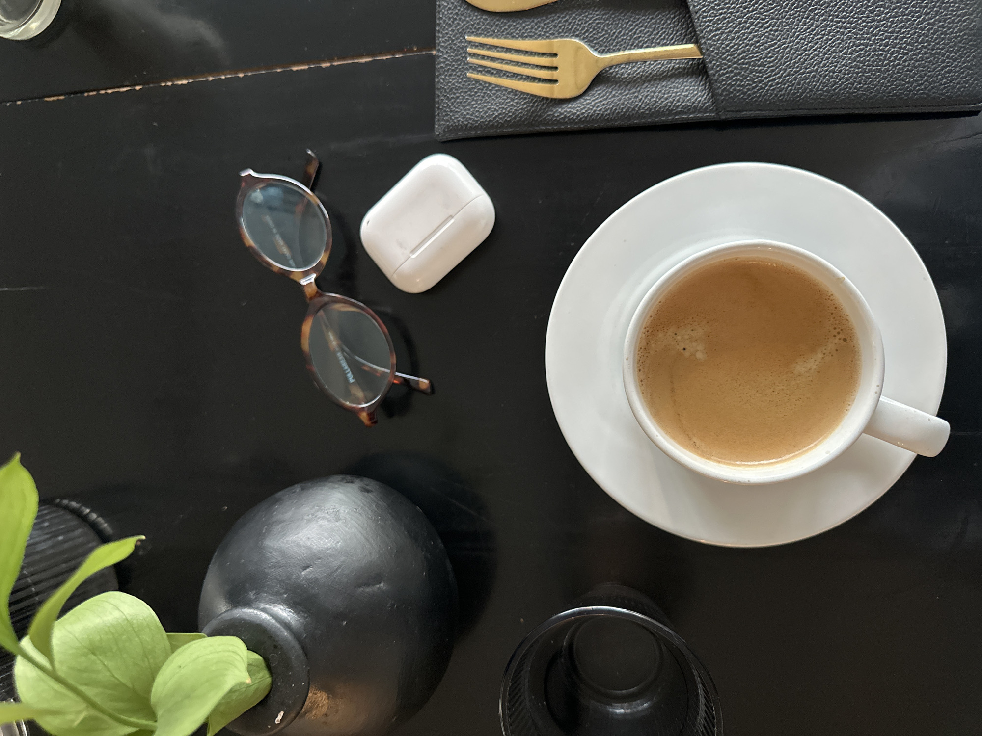 A high-angle, close-up shot captures a dark, textured wooden table in Kuta Selatan, Indonesia, set for a simple breakfast or break. In the foreground, a white ceramic cup of espresso sits on a matching saucer. To the left, a dark, textured vase holds a vibrant green plant. Adjacent to the plant is a dark, textured drinking glass. In the upper right quadrant, a pair of tortoise-shell glasses rests on the table, alongside a white wireless earbud case. To the right of the coffee cup, a black leather menu holder contains a single gold fork. The lighting appears to be natural, casting subtle shadows on the table surface, suggesting an indoor setting.