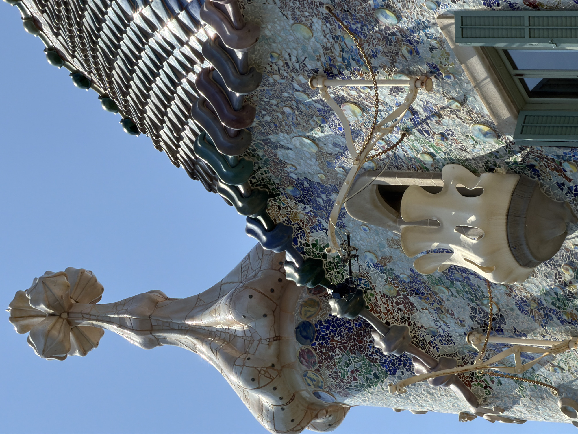 The image shows an architectural detail of Casa Batlló, located in Barcelona, Spain. The building's facade is covered in a vibrant mosaic of colorful tiles, predominantly blues and greens, with some circular iridescent elements. The roof features an undulating, scale-like pattern of glazed tiles in shades of green, brown, and purple. A distinctive, bone-shaped structure protrudes from the wall, and an ornate, flower-like finial crowns a conical turret. The architecture exhibits organic, flowing lines characteristic of Antoni Gaudí's style. A window with closed green shutters is visible in the lower right corner. The sky above is a clear, bright blue.