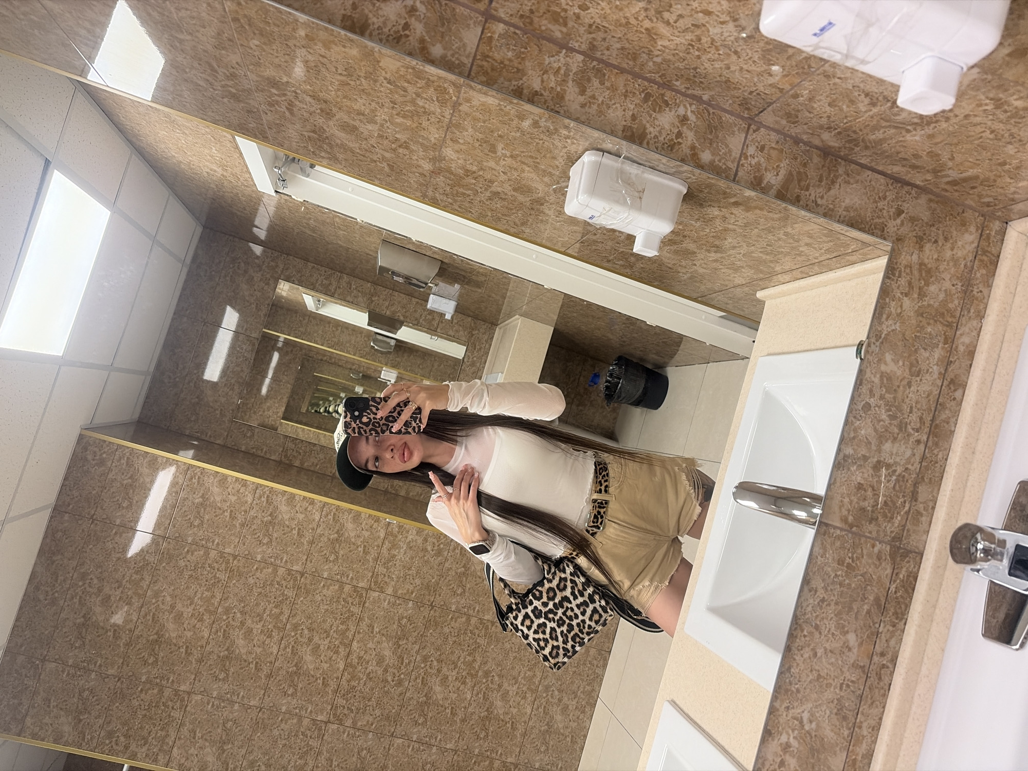 A young woman with long dark hair, wearing a white long-sleeved top, tan shorts, a leopard print belt, and a dark baseball cap, is taking a mirror selfie in a public restroom. She is holding a leopard print phone case and a leopard print purse. The restroom has tan marble tile walls, a white ceiling with fluorescent lighting, and a vanity with two sinks and chrome faucets. There are also soap dispensers and a trash can visible in the reflection. The image appears to be taken in Novosergievka, Russia.
