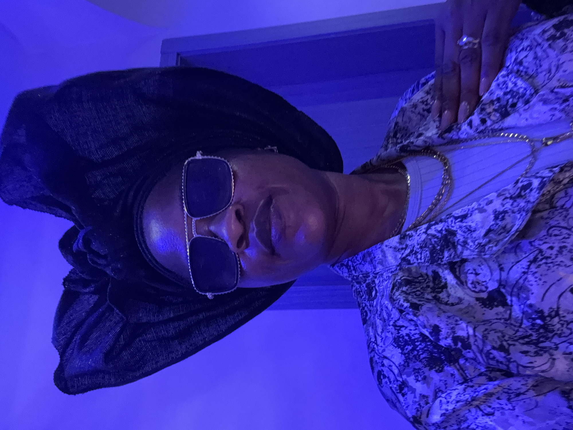 A woman wearing a black headwrap, sunglasses, and a patterned jacket is posing for a photo. She is indoors, with a blue light illuminating the scene. The woman is wearing multiple gold necklaces and a ring on her right hand. The background appears to be a wall.