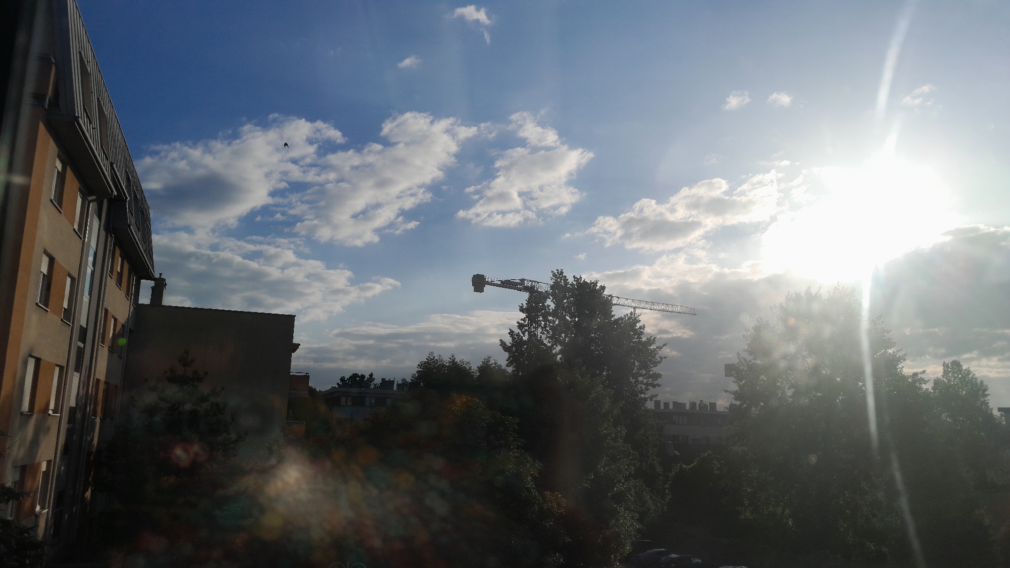 The image shows a sky filled with blue and scattered white clouds. The sun is bright and positioned in the upper right quadrant, casting strong lens flare and sunbeams downwards. A construction crane is visible in the middle distance, its boom extending horizontally across the frame. Silhouetted trees and buildings are in the foreground and middle ground, partially obscuring the view. The left side of the image features the side of a building with multiple windows. A small dark shape, possibly a bird, is visible in the upper left quadrant of the sky. The overall scene appears to be an urban or suburban landscape during the daytime, with a focus on the sky and construction elements.