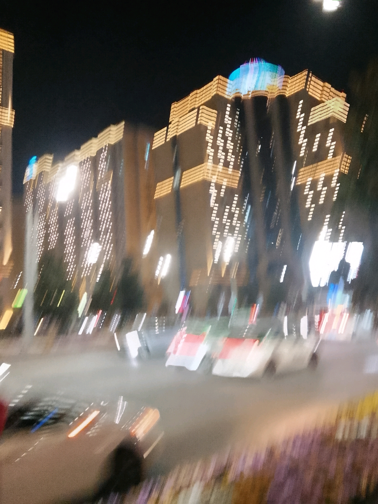 The image depicts a blurry, night-time scene in the city of Dushanbe, Tajikistan, likely in a modern, well-lit urban area. In the background, a large building, possibly a hotel or administrative center, is illuminated with rows of warm, golden lights and a distinctive blue and multicolored light display on its rooftop. The building's facade is adorned with a pattern of white lights that create a grid-like effect. In the foreground and mid-ground, streaks of light indicate moving vehicles and possibly streetlights or decorative lighting. Several cars are visible, their forms blurred by motion, with their headlights and taillights appearing as elongated streaks of white and orange. The road surface is visible, appearing wet or reflective. In the lower portion of the image, blurry elements suggest foliage or decorative flower beds, with streaks of purple and yellow colors. The overall impression is one of a vibrant, active urban environment at night, characterized by architectural illumination and vehicular movement.