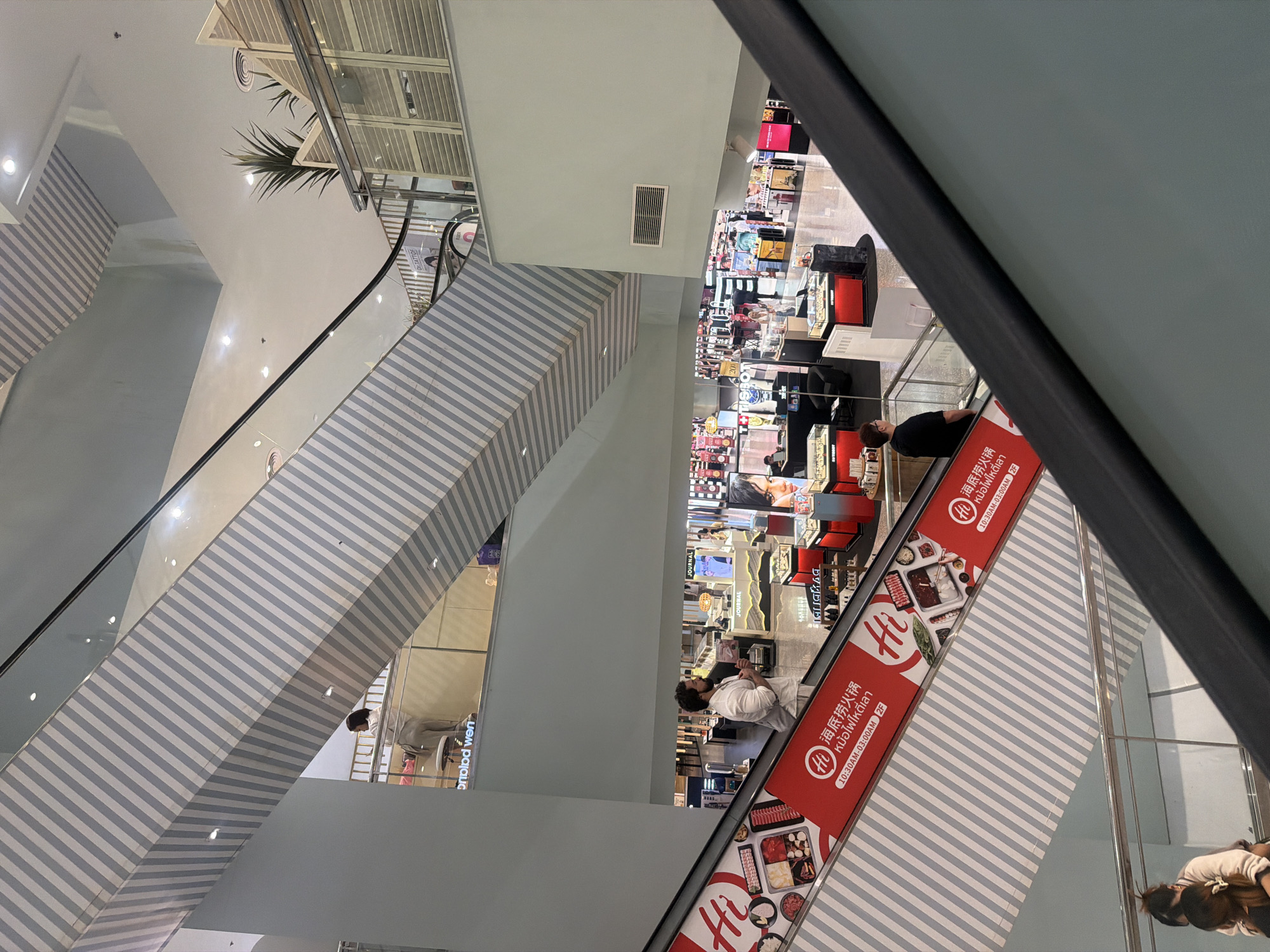 The image depicts a multi-level interior of a shopping mall. The foreground features a diagonal view of a light blue escalator with prominent red signage advertising "Hi" and what appears to be a food establishment, possibly hot pot, with text in both Chinese and Thai characters, including operating hours. Several individuals are visible on the escalators. On the escalator in the foreground, a man with dark hair and a beard is ascending, holding a book. Further up the escalator, two women are seen from behind.
The background reveals the architecture of the mall, with diagonal striped patterns on structural elements and balconies on upper levels. Shops with displays of merchandise, including jewelry or watches, are visible on lower floors, identified by signs like "JOURNAL" and "TISSOT". Plants are placed strategically on some levels. The lighting suggests an indoor environment with artificial illumination. The presence of Thai script on the signage indicates the location is likely in Thailand. Given the provided context, this scene is situated within a shopping mall in Wichit, Thailand.