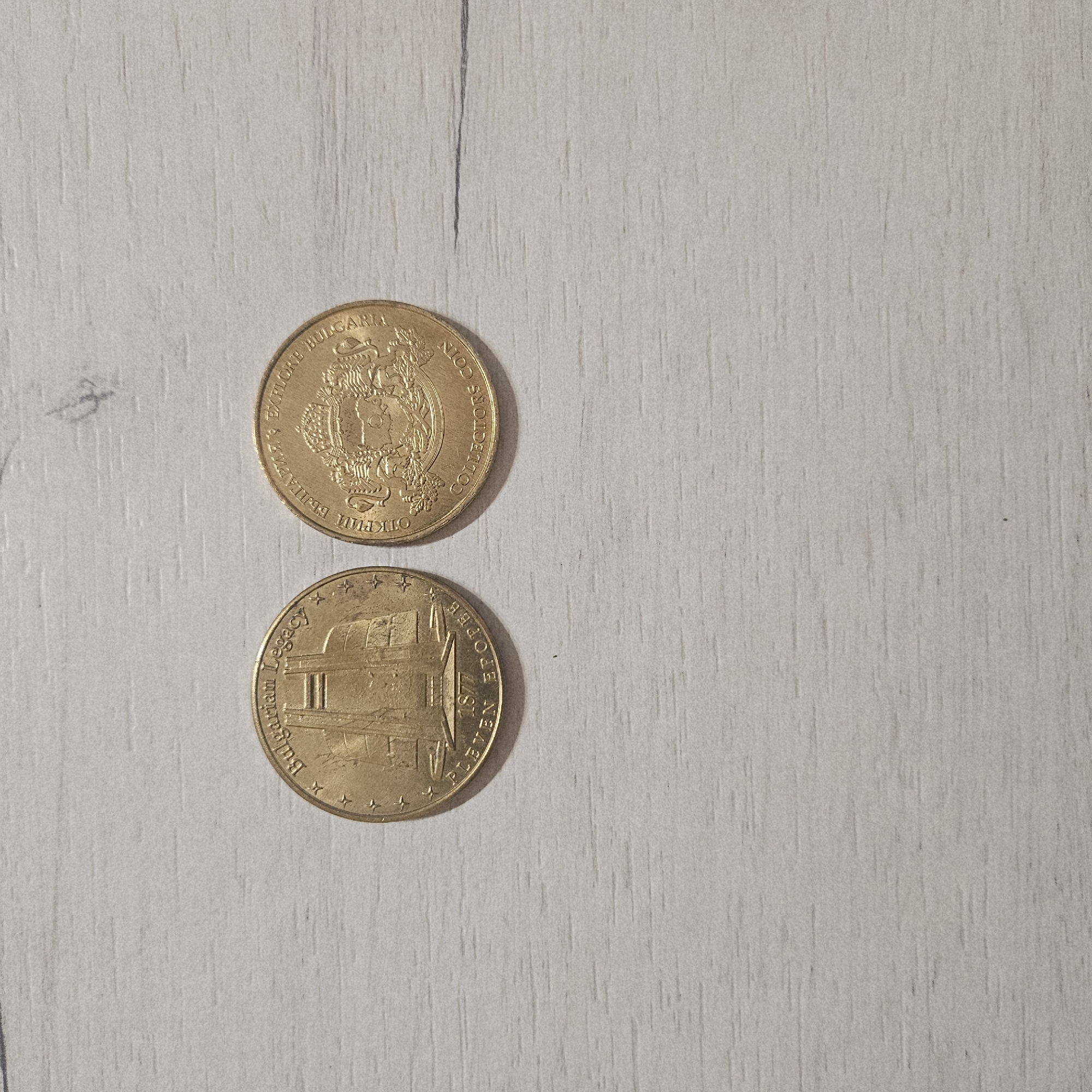 Two gold-colored commemorative coins are displayed on a light-colored, wood-grain surface.
The top coin features the Bulgarian coat of arms, which includes a crowned lion rampant within a shield, supported by two lions, and encircled by an oak wreath. Around the edge of this coin, the English text "EXPLORE BULGARIA" and "COLLECTORS COIN" is visible, alongside the Cyrillic text "ОТКРИЙ БЪЛГАРИЯ" (OTKRIY BALGARIYA), meaning "Discover Bulgaria."
The bottom coin depicts a historical building or monument, possibly related to the city of Pleven in Bulgaria. The visible English text on this coin reads "BULGARIAN LEGACY" and "PLEVEN 1877." There are four stars positioned on either side of the "BULGARIAN LEGACY" inscription.
The coins are stationary on the surface, which appears to be a table or countertop. The image is a close-up shot, focusing on the details of the coins against the textured background.