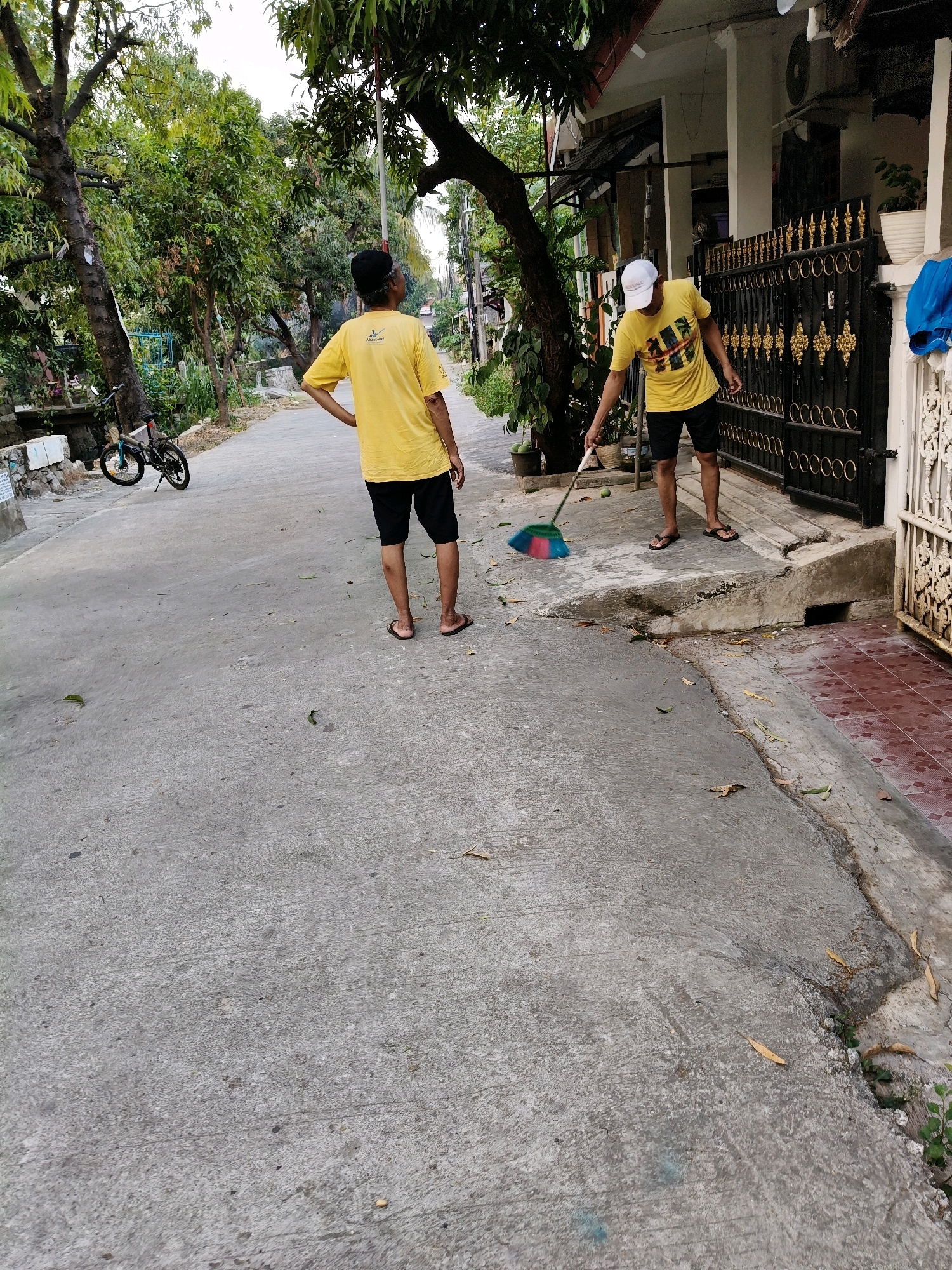 Two men are on a paved path in front of a residential building in Setiamekar, Indonesia. The man on the left, wearing a yellow t-shirt and black shorts with a black cap, stands with his hands on his hips, facing away from the camera. To his left, a bicycle is parked. The man on the right, in a yellow t-shirt, black shorts, and a white baseball cap, is sweeping the path with a broom that has a multi-colored head. The building behind them has a black metal gate and white ornate fencing. Trees and foliage are visible in the background.