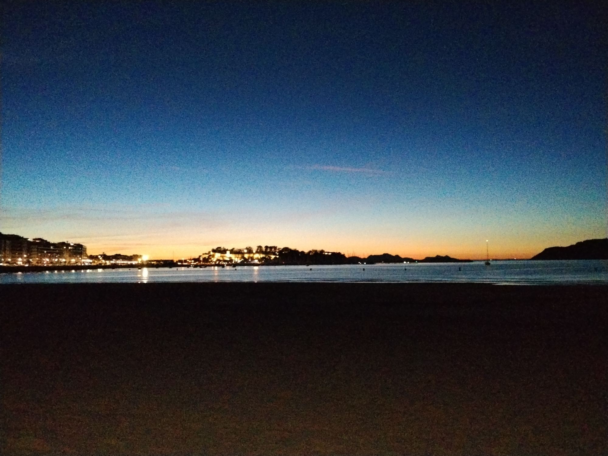 This image depicts a twilight view of the Bay of Baiona, Spain. The foreground is dominated by a dark sandy beach. Beyond the beach, the tranquil waters of the bay reflect the fading light and the distant city lights. On the horizon, the town of Baiona is visible, with its buildings illuminated and clustered together, suggesting a populated area. The sky transitions from a deep blue at the top to lighter shades of blue and orange near the horizon, indicating either sunrise or sunset. A few boats are present on the water, and the silhouette of a distant landmass or island can be seen on the right side of the frame. The overall atmosphere is peaceful and serene.