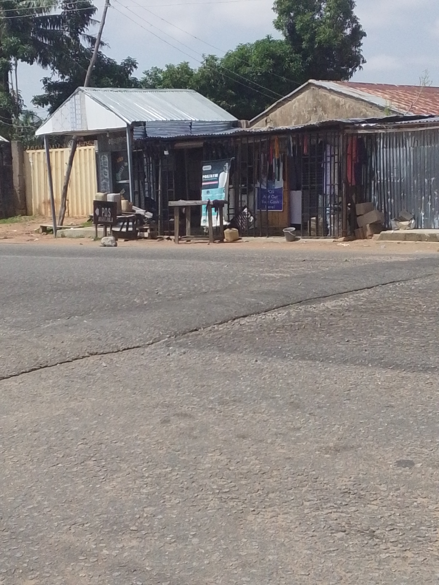 The image shows a street scene in Jalingo, Nigeria. A row of small shops with corrugated metal roofs and walls lines the side of a paved road. One shop has a sign that says "POS Available." Another has a poster that reads "POS Aid and Get Your Cash Here!". A few objects, possibly goods for sale, are visible through the metal bars of the shops, including what appears to be clothing. In front of the shops, there are several wooden tables and stools, as well as some buckets and what looks like a rock. The sky is overcast, suggesting it might be daytime, possibly late morning or early afternoon. The scene appears to be a typical commercial area in a Nigerian town, with a focus on small businesses and services. There is no clear indication of any particular event or activity other than the general operation of these small shops.