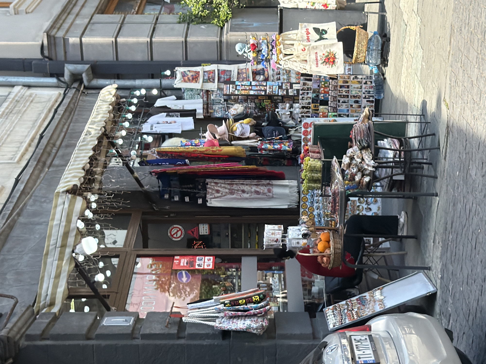 The image depicts a street-level view of a souvenir shop in Tbilisi, Georgia. The storefront features a striped awning with string lights. Various goods are displayed outside, including bags, clothing like t-shirts, hats, scarves, and an array of small souvenir items. A person wearing a red top and black pants is seated on a chair behind a table laden with fruit, such as oranges, and other merchandise. A silver car is parked on the left side of the frame, its license plate visible. The scene is set on a cobblestone street with sunlight casting shadows.