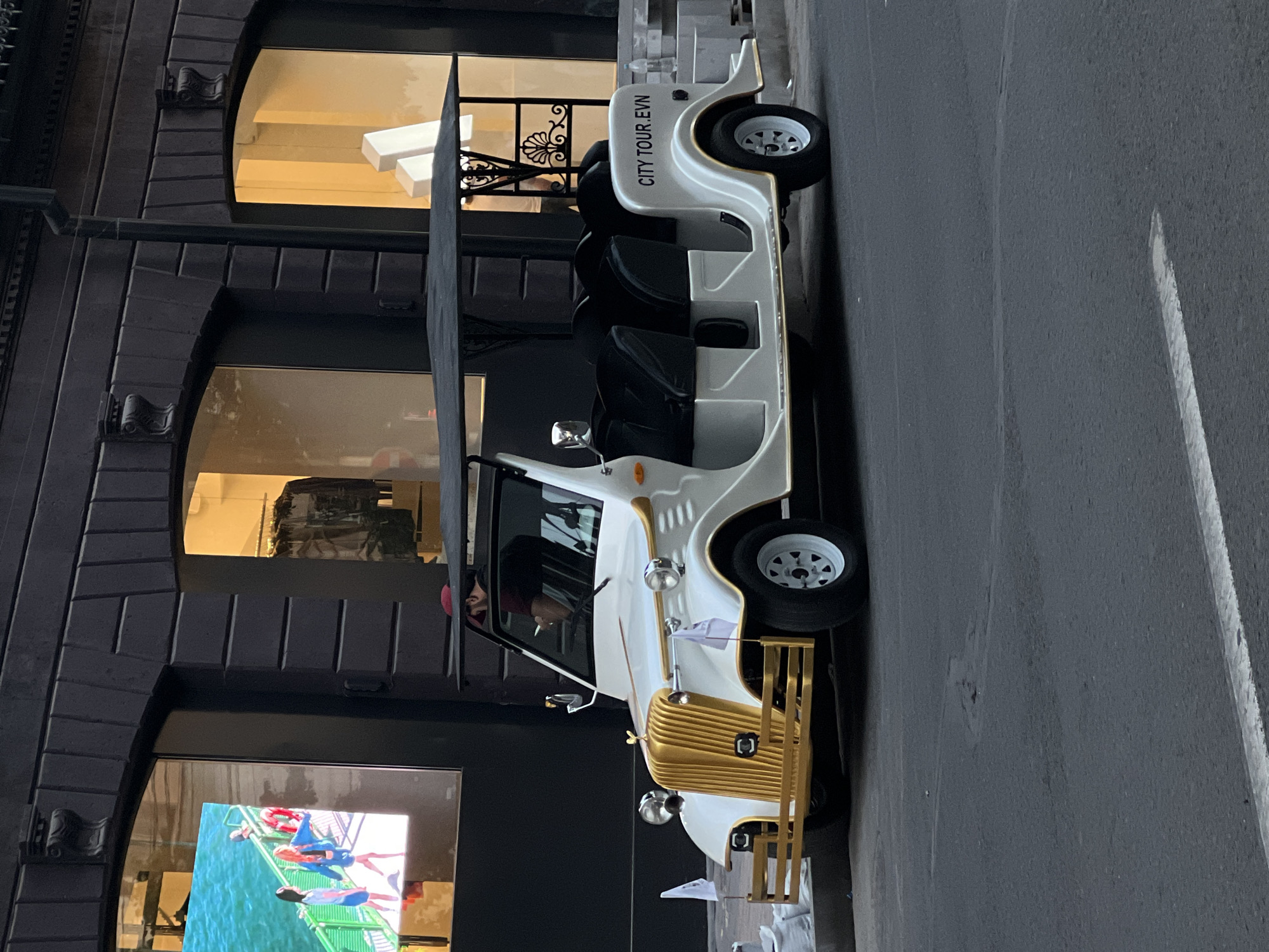 A white and gold city tour vehicle is parked on a gray asphalt street next to a dark building with large arched windows. The building has detailed stone or brickwork around the windows. One window displays mannequins, while another shows a video of people swimming. The vehicle is labeled "CITY TOUR.EVN" on its rear panel. A person can be seen in the driver's seat.