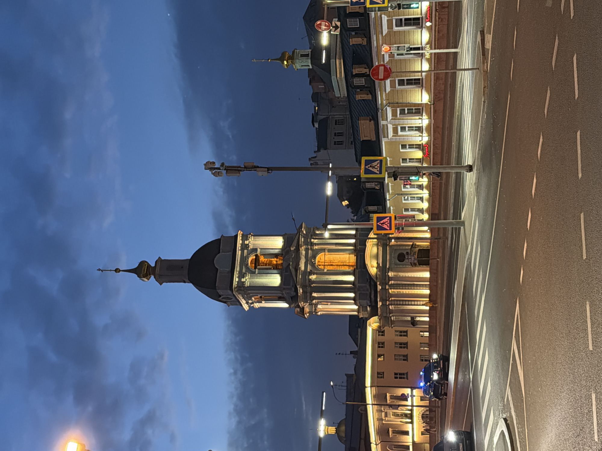 The image shows a night view of the Moscow Theological Academy and its bell tower, part of the Trinity Lavra of St. Sergius complex, located in Sergiyev Posad, near Moscow, Russia. The bell tower is illuminated with warm lights, highlighting its architectural details. In the foreground, a street with a pedestrian crossing is visible. A police car with flashing blue lights is driving across the intersection. The sky is dark blue with some clouds. Several traffic signs and streetlights are also present.