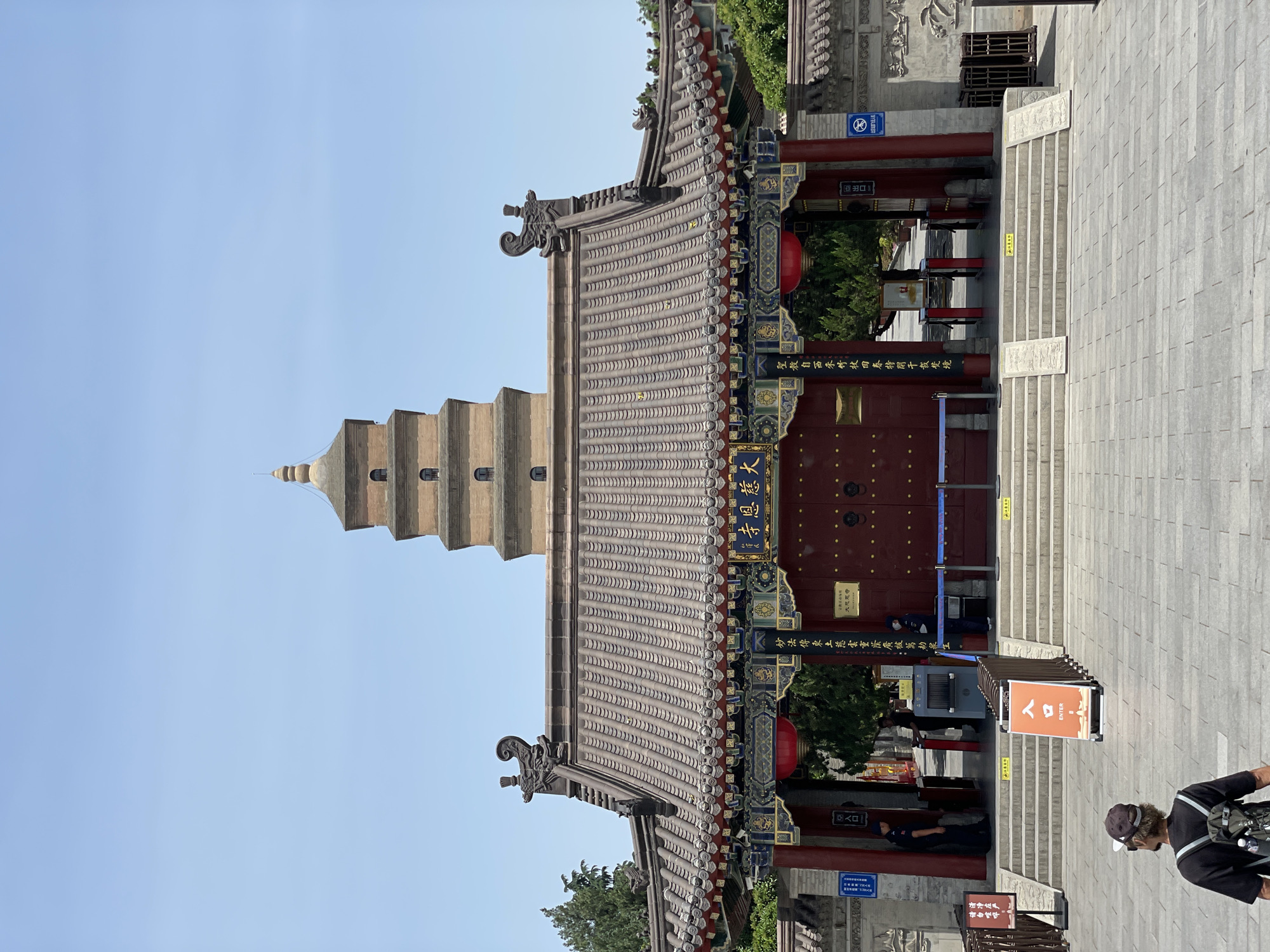 The image shows the Da Cien Temple in Yanta District, China. In the foreground is a traditional Chinese-style gate with a red door and ornate roof. A pagoda-like structure is visible behind the gate, towards the left. There are steps leading up to the gate. A sign marked "ENTER" in both English and Chinese is placed near the steps. A person with a backpack is partly visible in the bottom right corner. The sky is clear and blue.