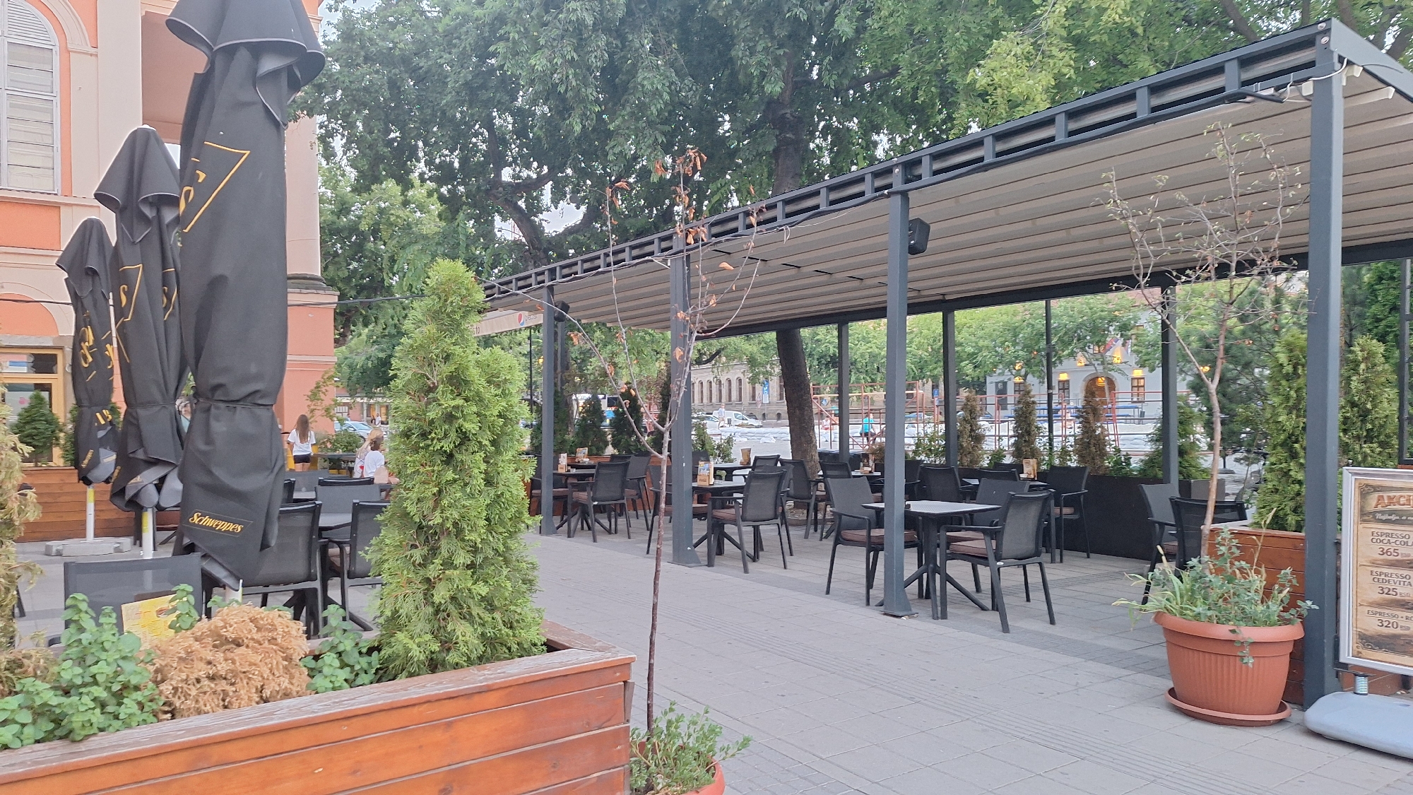 An outdoor seating area of a cafe is pictured in Sombor, Serbia. The area is covered by a modern, gray metal awning with a white retractable roof. Several tables and chairs are arranged on a paved patio. To the left, a row of tall, black, closed umbrellas with yellow branding are displayed, one of which clearly shows the word "Schweppes." Greenery, including small trees and shrubs in wooden planters, is strategically placed throughout the seating area. In the background, blurred figures of people can be seen, and buildings with architectural details characteristic of European cities are visible. A sign with menu items and prices is posted on a stand on the right side of the image. The overall scene depicts a well-maintained and inviting outdoor dining space on a cloudy day.