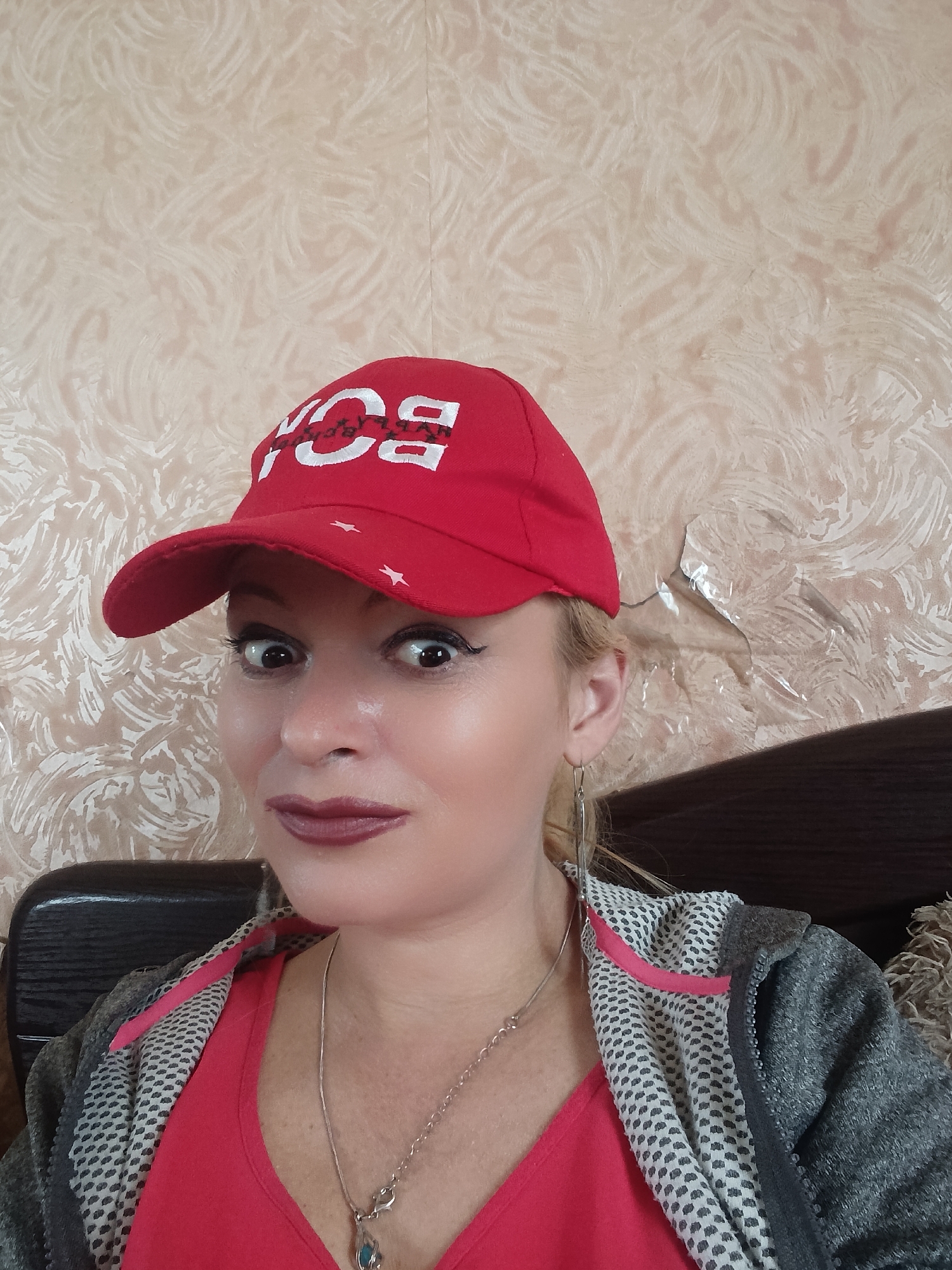 A woman is pictured from the chest up, wearing a red baseball cap with white lettering that reads "BOY", a red v-neck shirt, and a grey zip-up hoodie. She has blonde hair, dark eyeliner, and dark lipstick. She is wearing a silver necklace with a blue pendant and dangling silver earrings. The background is a beige patterned wallpaper. The image appears to be taken indoors, possibly in an apartment, in the city of Chernihiv, Ukraine.