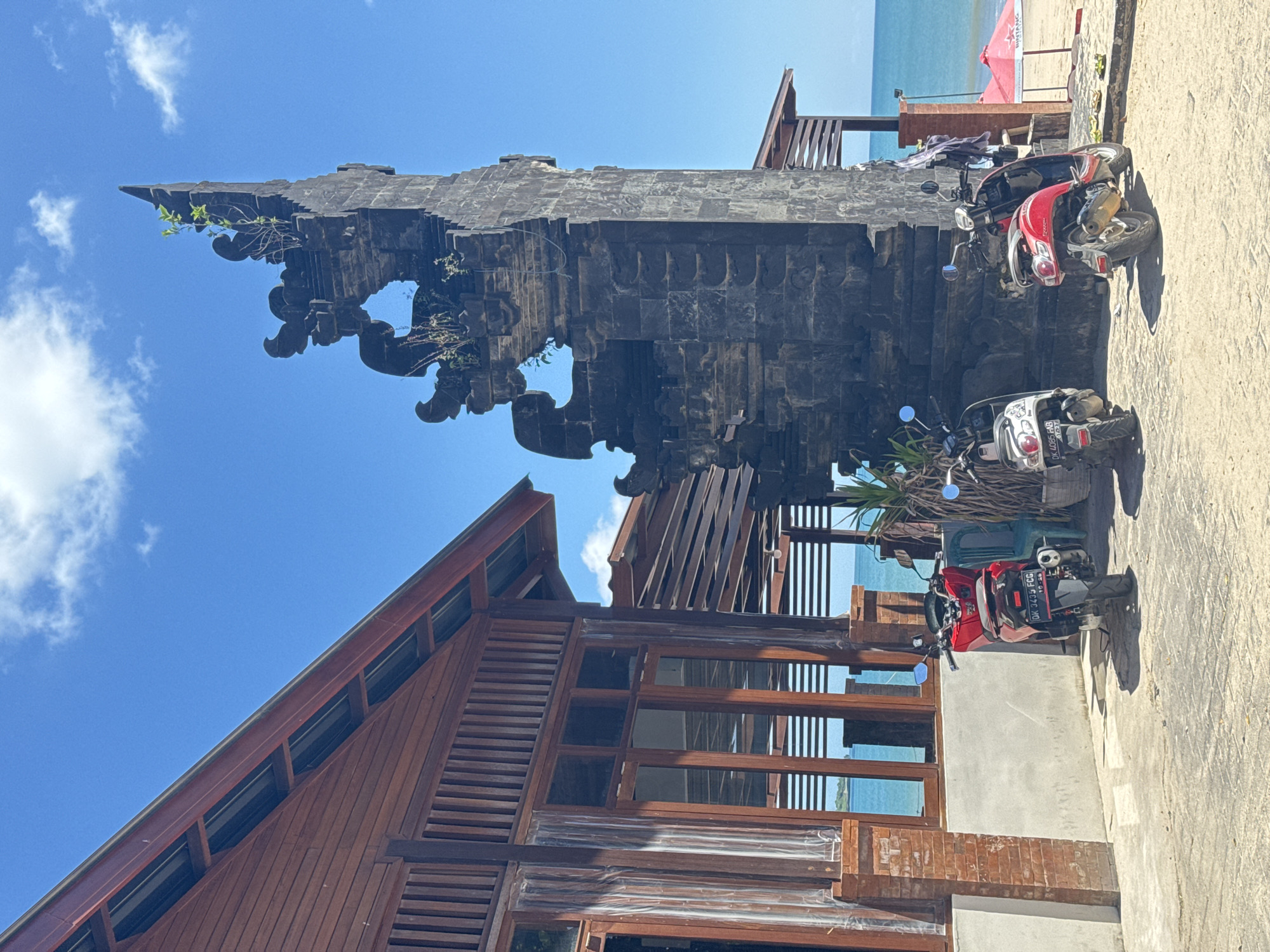 The image depicts a scene on what appears to be a beach in an unidentified city. A traditional Balinese stone gate structure stands prominently, featuring intricate carvings and weathered texture. The gate is partially overgrown with vegetation. Adjacent to the gate is a wooden building with a steep, A-frame roof and numerous windows. Several motorcycles are parked nearby on a concrete or paved surface. The background reveals a sandy beach and the ocean under a clear, blue sky with scattered clouds. A red umbrella or canopy is visible in the distance on the beach.