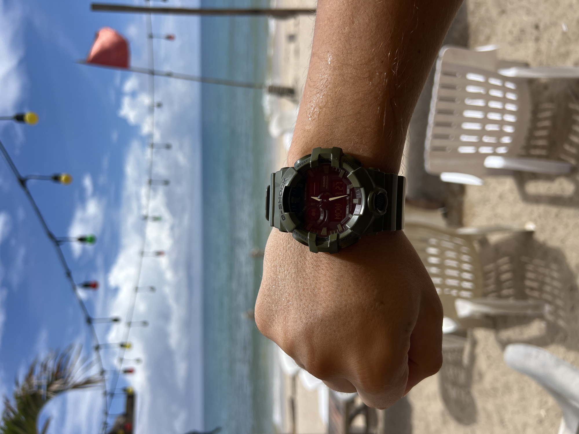 A person's left wrist, adorned with a green G-Shock watch, is prominently displayed in the foreground. The watch face shows the time as 12:27 and the day as Monday. The background depicts a beach scene with the ocean, a sandy shore, white plastic lounge chairs, and a string of colorful lights overhead. A red flag waves in the distance against a bright blue sky dotted with white clouds. The overall atmosphere suggests a tropical vacation setting.