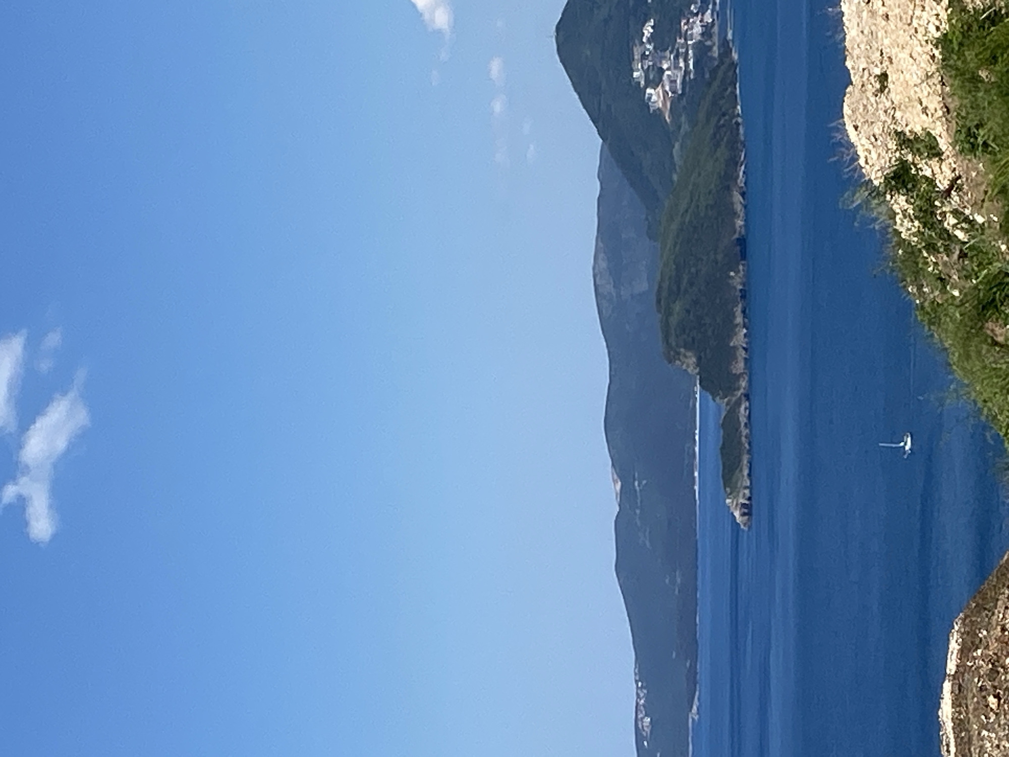 The image depicts a coastal scene in Blizikuće, Montenegro. The sky is clear and blue with a few scattered clouds. The Adriatic Sea is a deep blue, with a single sailboat visible in the distance. A series of mountainous islands, covered with green vegetation, rise from the sea. A cliff face with vegetation is in the foreground. Buildings dot the landscape on top of one of the mountains.