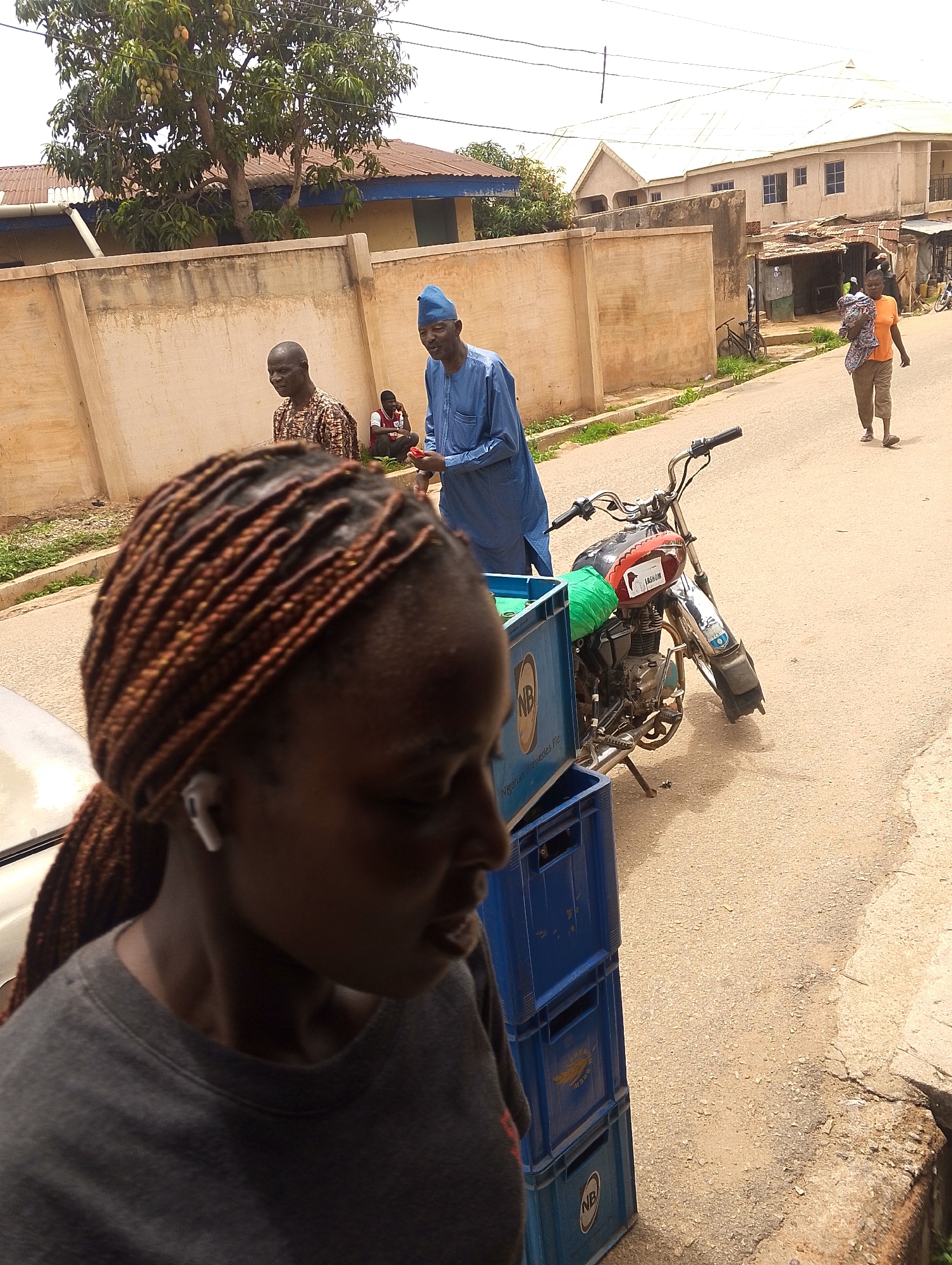 The image shows a street scene in Jos, Nigeria. In the foreground, a woman with braided hair and an earbud is partially visible. Behind her, a stack of blue "NB" (Nigerian Breweries) crates stands next to a parked motorcycle. Further back, two men are standing by a wall, with a seated person nearby. A woman carrying something on her back walks away in the distance. The street is paved and flanked by buildings and walls. A tree is visible above the wall on the left.