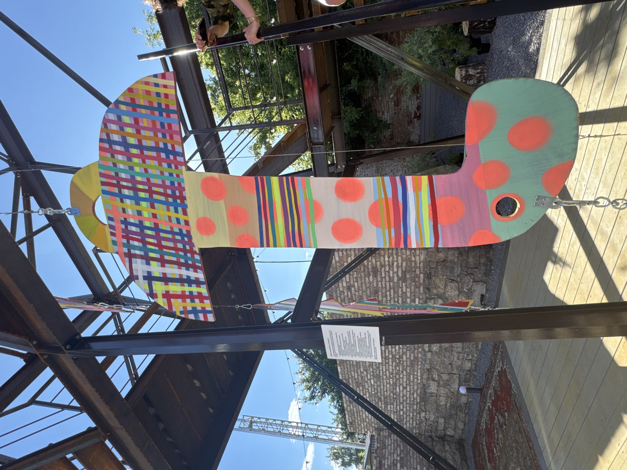 An outdoor, vertical, colorful, stylized art installation, resembling a mushroom or a sock, hangs from a metal structure under a bright blue sky with scattered clouds. The installation features a patterned top with plaid-like designs in various bright colors, a long stem with bands of multi-colored stripes and large orange polka dots, and a bulbous, green base adorned with orange polka dots. A circular cutout is visible on the lower part of the base. The art piece is suspended by metal chains and hooks. In the background, a brick wall is visible, along with a wooden deck, an ornate rug on the ground, and some greenery. To the right, a person's hands are seen reaching towards the installation, holding what appears to be a phone or camera. To the left, a white sign with text is attached to a black metal support post. The overall scene suggests an art exhibition or public display in an urban outdoor setting. This artwork is located at the Yelets Museum of Local Lore in the city of Yelets, Russia.