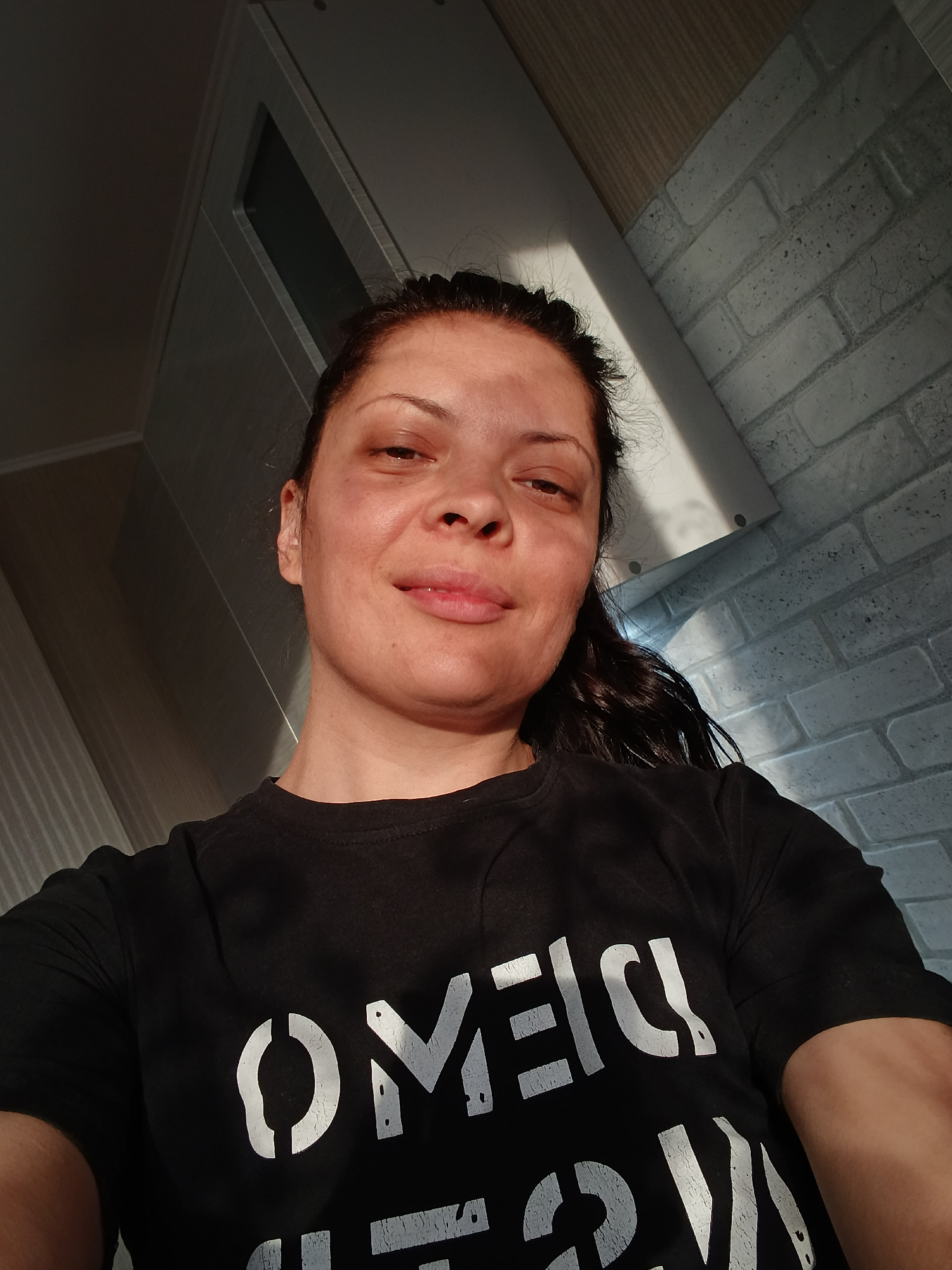 An indoor, low-angle selfie captures a woman, likely an adult, looking towards the camera with a calm, subtle smile. Her dark hair is pulled back, and she is wearing a black t-shirt featuring large, white, distressed, stencil-like letters. Visible portions of the text include "OMEI" or "OMEL," strongly suggesting the word "OMEGA" or "ОМЕГА."
Strong, natural light, indicative of daytime (possibly morning or late afternoon), illuminates the right side of her face and body, creating pronounced highlights and shadows.
The background reveals an interior setting, likely a home in Saratov, Russia. To the upper left, a light grey cabinet or shelf unit with a dark rectangular insert is visible. The wall behind and to the woman's right features a decorative light grey brick pattern, while other wall and ceiling sections appear plain light grey or white. The camera's low perspective emphasizes the upper parts of the room.