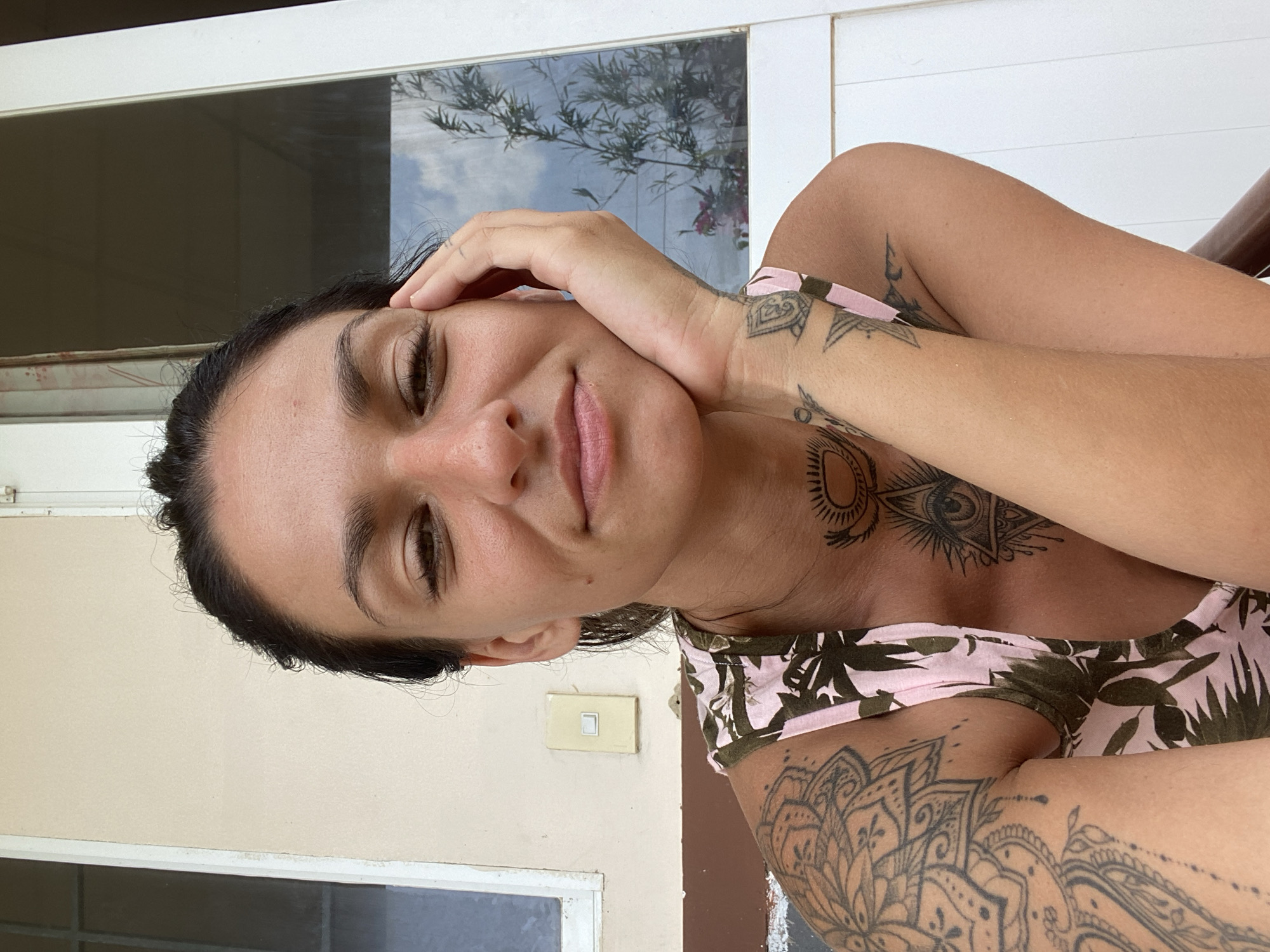 A young woman with dark hair and tattoos on her arms and chest is pictured in a close-up selfie. She is resting her chin on her hand and smiling. The background appears to be outdoors with a light-colored wall, a window with a reflection of trees and sky, and white trim. A light switch is visible on the wall. The woman is wearing a pink and green floral tank top. This image was taken in Mũi Né, Vietnam, likely on a porch or veranda.