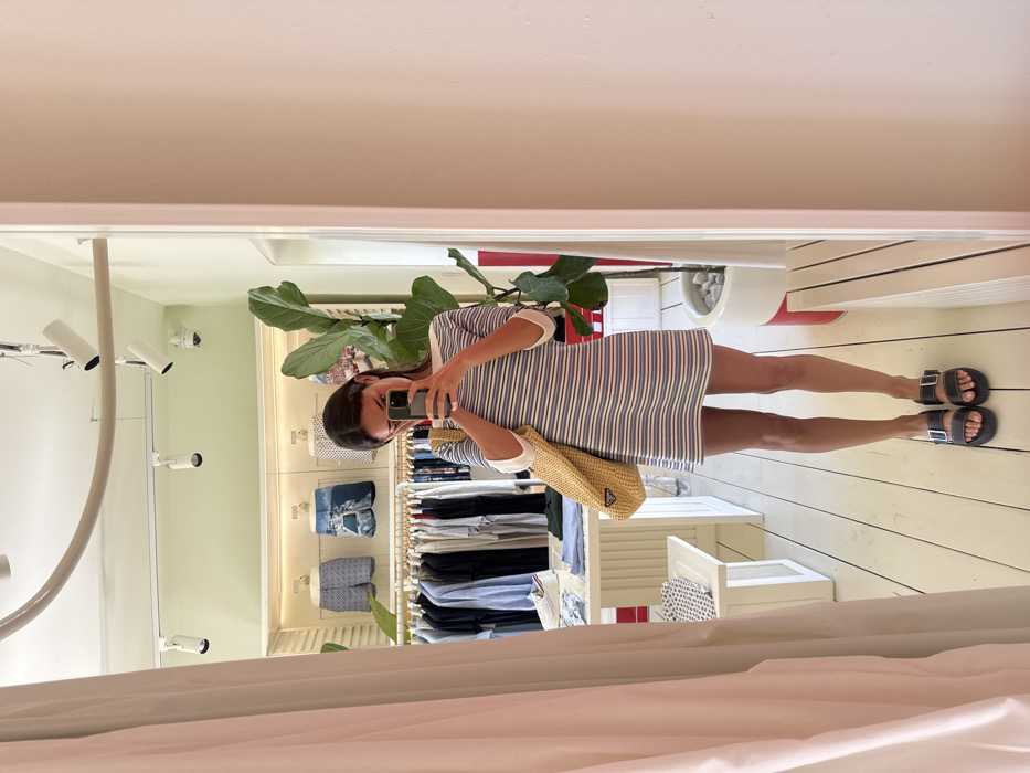 The image depicts a female individual taking a mirror selfie inside a clothing store in Marina Maroma, Mexico. The subject, viewed from the waist up in a frontal pose, wears a short-sleeved, boat-neck dress with horizontal blue and white stripes and white sleeve cuffs. She holds a dark smartphone in both hands at chest level, positioning it to capture her reflection. A large, light-colored woven straw bag with a dark triangular logo, resembling Prada, is slung over her left shoulder. She wears dark, two-strap sandals on her feet.
The environment, visible in the mirror's reflection, includes light-colored wooden floorboards, light green walls, and a white ceiling with a track lighting system featuring multiple white spotlights. A curved white conduit and a dome-style security camera are also present on the ceiling. To the left, a white display table holds folded clothing items, with a clothing rack behind it displaying various garments on hangers. Above this rack, three light-colored display units showcase folded swim shorts. A large green potted plant, with broad leaves, stands in a white pot with a red stripe to the right of the main display area. A light pink wall section and a light pink curtain partially frame the scene on the right and left, respectively.