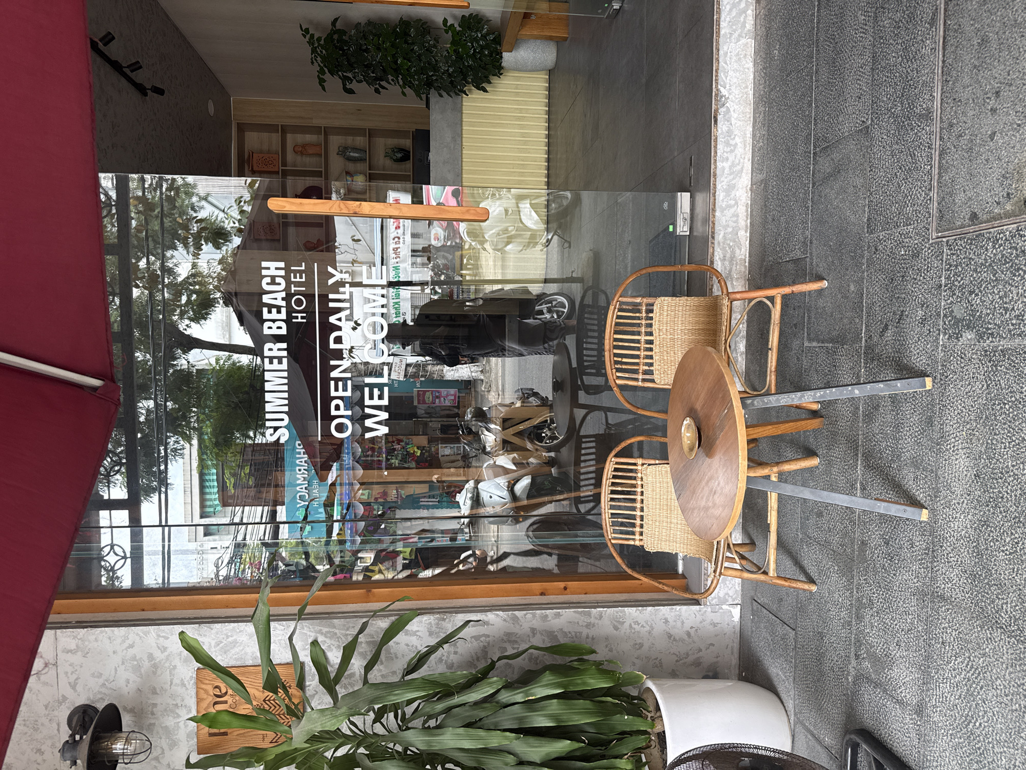 This image captures the exterior and reflected street scene of the Summer Beach Hotel in Đà Nẵng, Vietnam, specifically located at 20 Phan Liêm, Ngũ Hành Sơn.
In the foreground, a paved sidewalk features a small outdoor seating arrangement: two rattan chairs with woven seats flank a round, wooden-topped table with dark metal legs. A small, golden-brown ceramic dish rests on the table. To the far left, a large potted plant with long green leaves sits in a white cylindrical pot, with a portion of a black pedestal fan visible beside it. The sidewalk is composed of dark grey, textured square pavers.
The midground is dominated by a large glass entrance door for the "SUMMER BEACH HOTEL," with "OPEN DAILY" and "WELCOME" inscribed in white, capitalized letters. The glass reflects the street scene opposite the hotel, including several motorbikes (at least two light-colored scooters and one darker bike) and a person wearing a helmet, possibly on a motorbike, obscured by the reflections. Overhead electrical wires are visible in the reflection, along with the façades of buildings across the street, one of which appears to be a "PHARMACY" (reflected as "YOAMRAHP"). The door is framed by wooden uprights, and a red awning extends over the entrance from above. To the left of the door, an industrial-style wall lamp with a caged bulb is mounted on a light grey, textured wall, below which a wooden sign, partially reading "One Coffee," is affixed.
Through the clear glass door, the hotel's interior reception area is visible. A reception desk with a textured yellow lower panel and a dark grey top stands to the right. Behind it, a wooden shelving unit displays various decorative objects, including vases, statues, and lanterns. Another potted plant is situated inside to the right of the reception desk. The interior floor is a dark grey. The ceiling above the reception area features black track lighting.