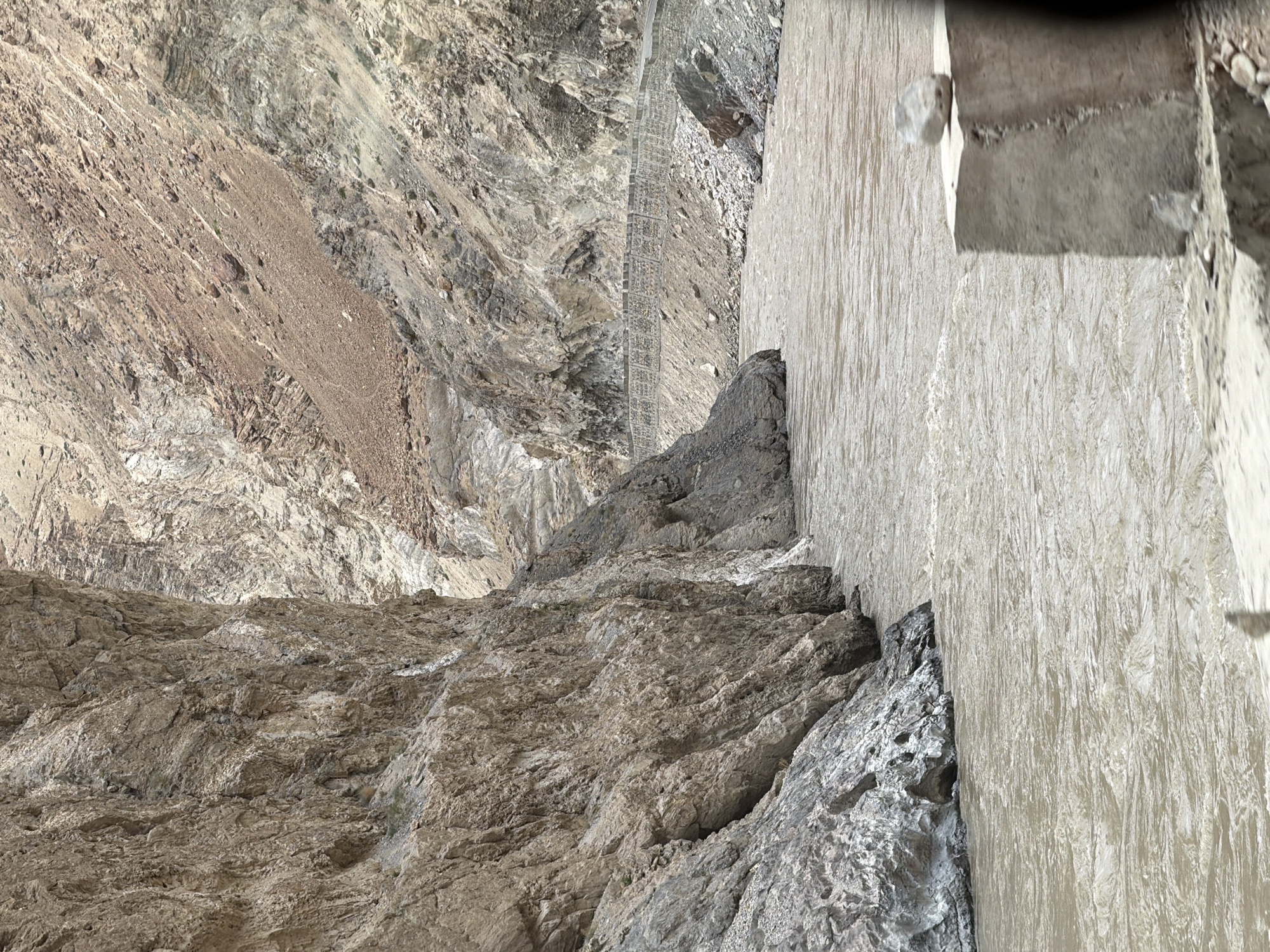 A wide, muddy river flows through a narrow canyon. Steep, rocky cliffs rise on either side of the river, with a road visible carved into the side of the right cliff. The terrain is arid and mountainous, with sparse vegetation. The water appears to be fast-moving. The scene suggests a remote, natural landscape. This location is in Stongde, India.