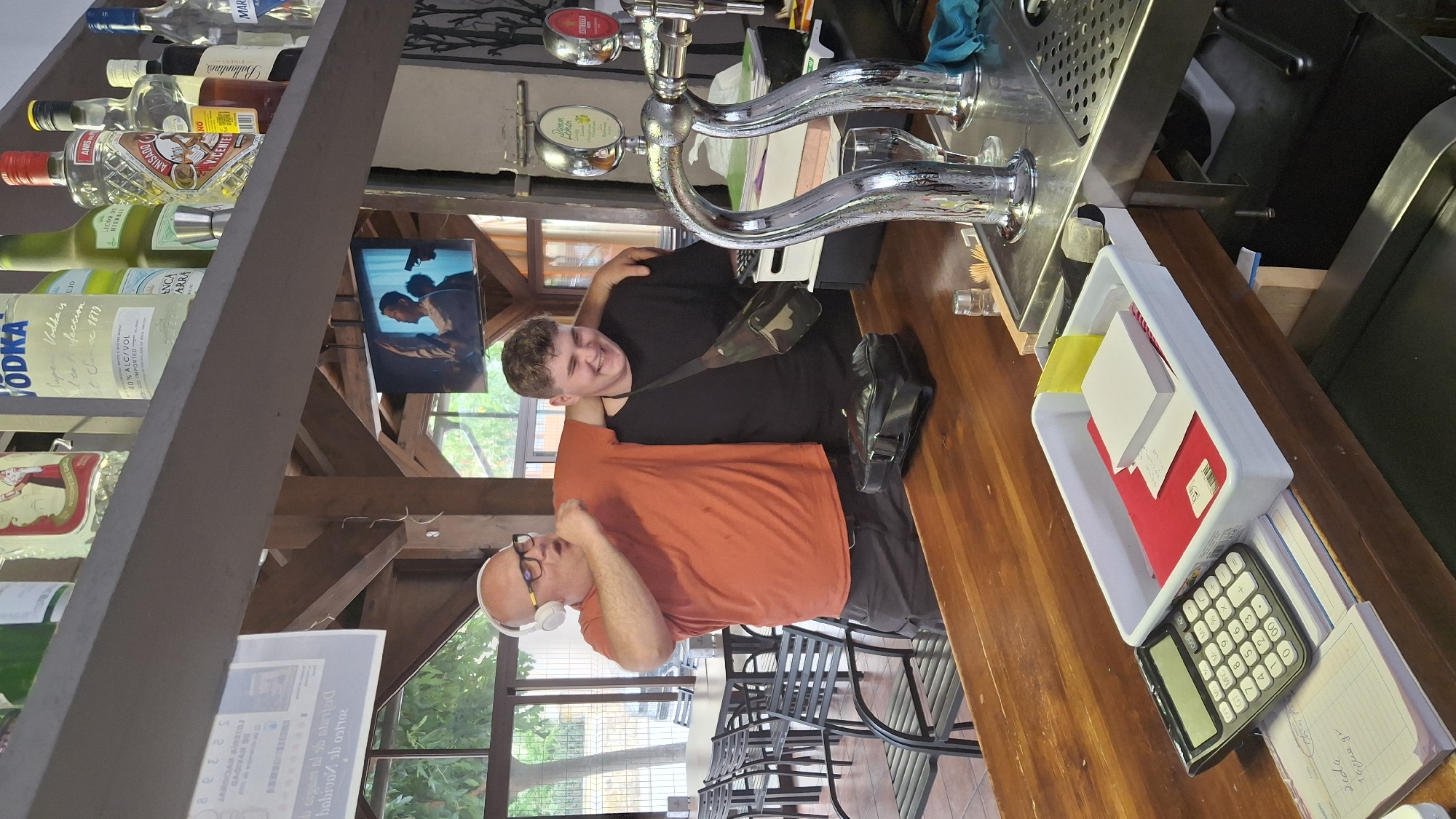A man wearing headphones and an orange t-shirt is dancing and singing while a younger man in a black t-shirt with a crossbody bag laughs beside him. They are standing behind a wooden bar counter. On the counter, there is a calculator, a white plastic tray containing papers and sticky notes, and a black bag. In the background, a television screen displays a scene from a movie. The bar appears to be an indoor-outdoor establishment, with tables and chairs visible through large windows and wooden beams supporting the roof. The establishment is likely located in La Fresneda, Spain, as indicated by a lottery ticket visible on the counter with Spanish text and a seal that appears to be from Spain. Bottles of alcohol are displayed on shelves above the bar.