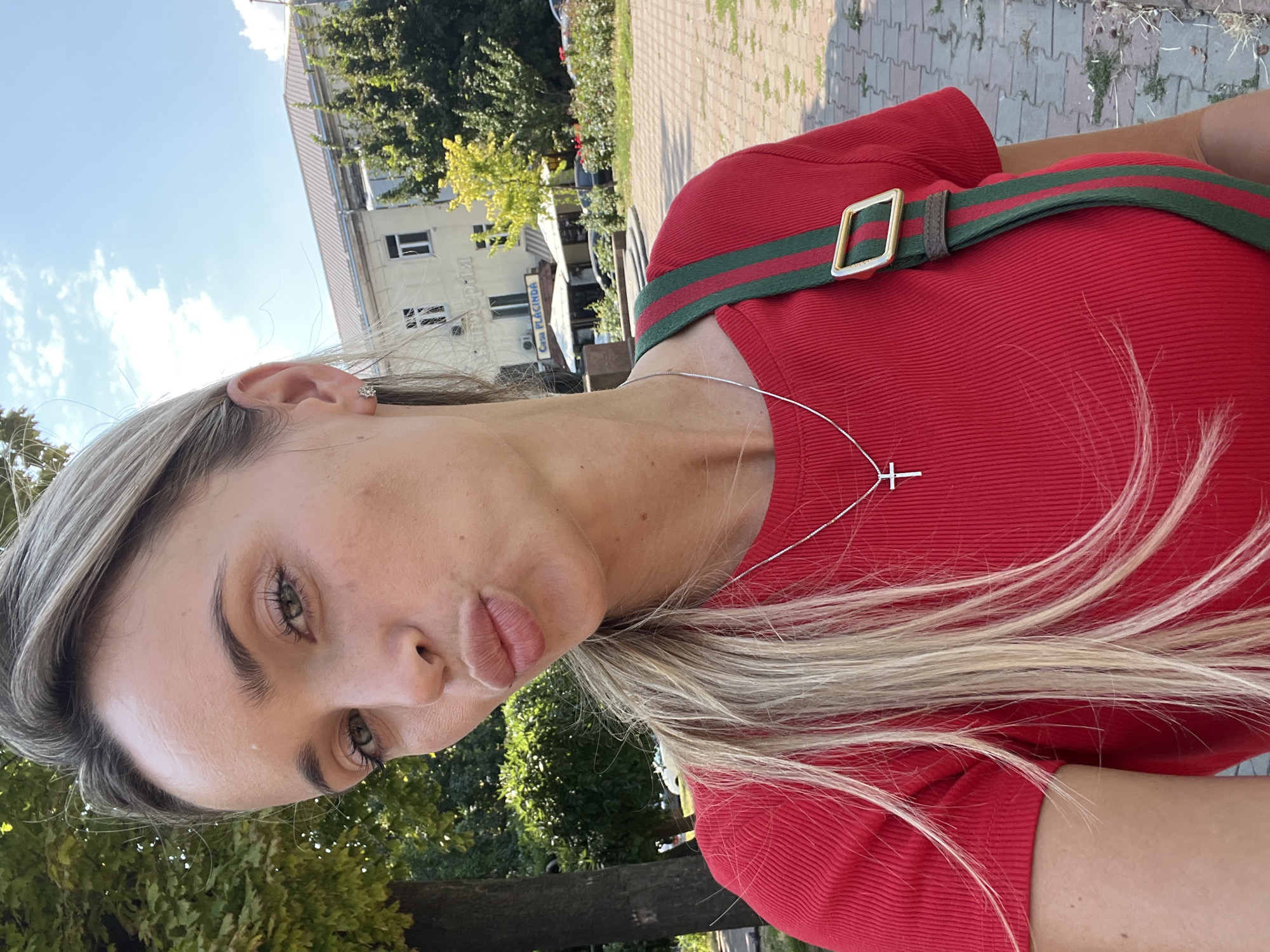 A blonde woman with fair skin and green eyes takes a selfie outdoors, puckering her lips. She is wearing a red ribbed t-shirt with a silver cross pendant on a chain. A green and red striped strap with a gold buckle is visible across her chest. Her hair is blonde and falls past her shoulders. In the background, a building with a tiled roof and arched windows stands behind trees and a paved walkway. The building has signage that appears to be in Cyrillic and English, including the name "CASA PLACINDA." The overall setting suggests a sunny day in a public area of Odesa, Ukraine.