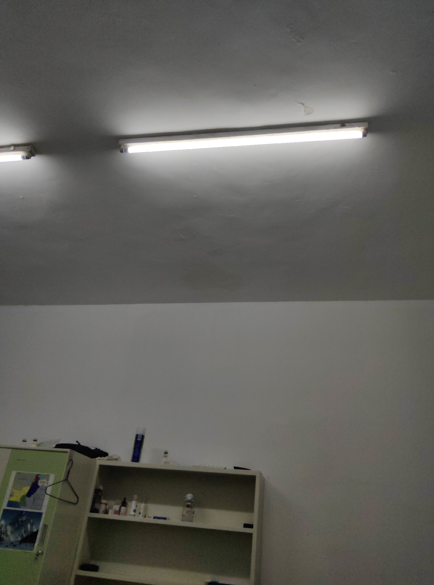 This is a dimly lit, indoor photograph of what appears to be a room or storage area. The upper portion of the image shows a white ceiling with two long fluorescent light fixtures, both illuminated. The ceiling has some minor imperfections or discoloration. The lower portion of the image shows a white wall. To the left, a light green cabinet or closet is visible, adorned with several posters or pictures. A black clothes hanger is hanging from the side of this cabinet. To the right of the cabinet is a white shelving unit, partially filled with various items, including bottles, a blue marker, and other indistinct objects. The overall scene suggests a functional, possibly utilitarian space. There are no people visible in the image. The location is stated to be in the Chongqing High-tech Zone, China.