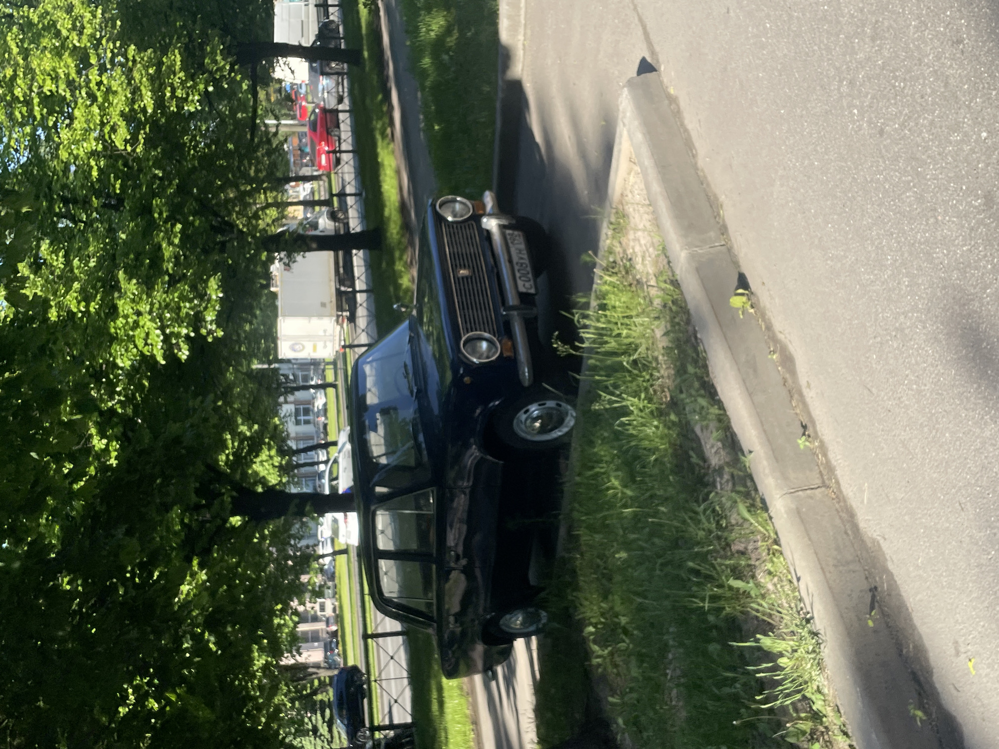 The image captures an outdoor scene featuring a dark-colored, older model sedan, resembling a Lada or Fiat 124 variant, parked on the side of a road. The car is positioned with its front bumper slightly angled towards the viewer, resting partially on a strip of grass adjacent to a concrete curb. Beyond the curb is an asphalt road surface.
To the left of the car, lush green trees with dense foliage fill a significant portion of the frame, suggesting a spring or summer season. Bright sunlight illuminates the entire scene, casting clear shadows and highlighting the vibrant green leaves. In the background, visible through gaps in the trees and beyond a black metal fence, are other parked vehicles, including white trucks and a red car, along with hints of buildings.
No people are visible in the frame, and the scene depicts a static moment with no discernible activity taking place, other than the car being parked. The time of day is clearly daytime, characterized by the strong, direct sunlight and distinct shadows. The weather appears clear and sunny.
A notable detail is the license plate on the front of the dark car, which prominently displays the alphanumeric sequence "C008YH 198". The "198" region code on the license plate suggests a potential location in St. Petersburg, Russia, which is consistent with the type of vehicle commonly found there. The overall scene conveys a calm, uneventful roadside setting.