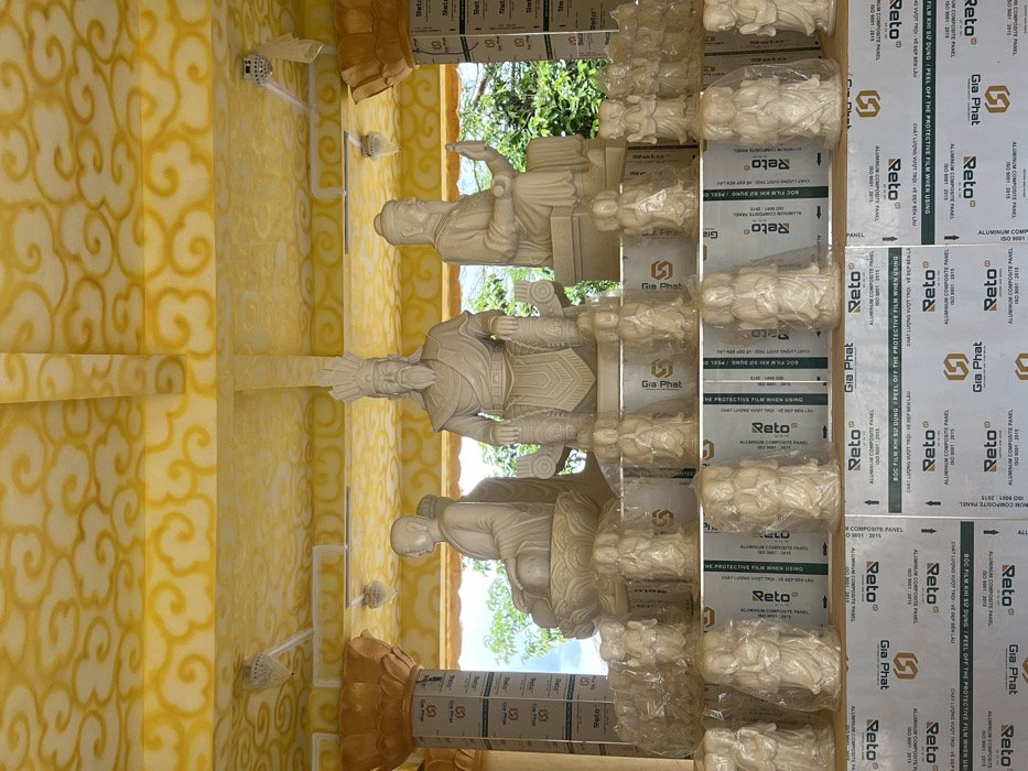 The image depicts an open-air shrine or temple structure located in Đăk Mar, Vietnam. The ceiling is painted yellow with a repeating white cloud motif and features several white ceiling fans. Ornate, gold-colored pillars with lotus-like capitals, partially covered by protective film, frame the scene on the left and right.
Three large, light-colored carved stone statues are positioned centrally. On the left, a seated figure with a bald head and monastic robes meditates on a lotus pedestal. In the center, a prominent, bearded figure with an ornate headdress and ceremonial attire is seated on a throne. To the right, another bearded, robed figure with a hat sits with one hand raised.
Below these main statues, multiple rows of smaller, light-colored carved figures, many of which are individually wrapped in clear plastic, are displayed. These figures typically show individuals with hands clasped. They are arranged on platforms constructed from stacked rectangular panels. These panels are silver-colored aluminum composite panels, branded "Gia Phat" and "Reto," with text in both Vietnamese and English indicating "ALUMINIUM COMPOSITE PANEL" and instructions regarding protective film removal. Green foliage and trees are visible in the background, suggesting an outdoor or semi-outdoor setting.