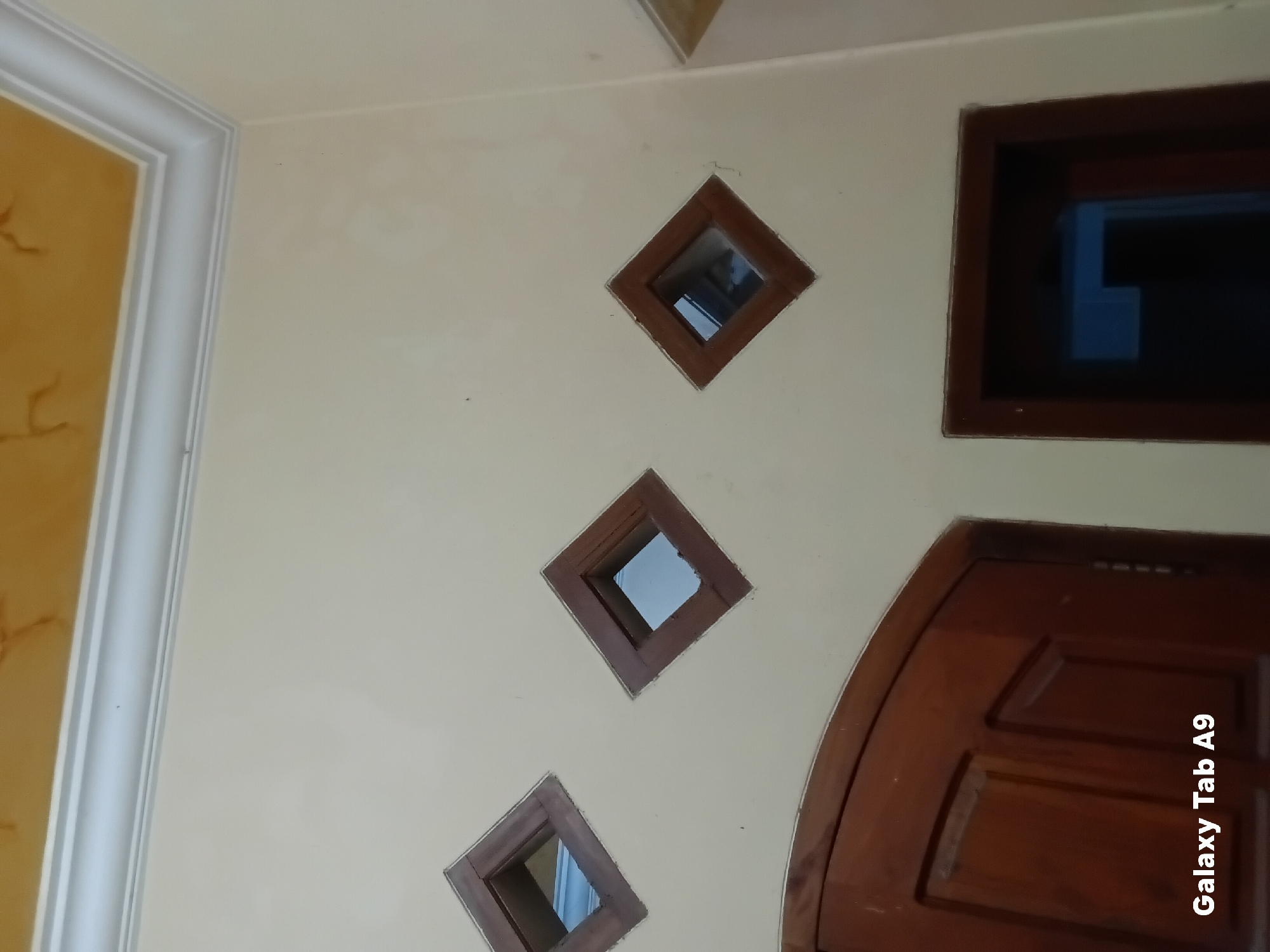 The image depicts an interior wall with cream-colored paint. Along the top of the wall, a white decorative cornice runs horizontally, and above it, a yellow ceiling with brown marbling is visible. Three diamond-shaped ventilation openings are set into the wall, each framed with dark wood. The openings reveal a darker interior space. In the lower left of the image, a portion of a dark wooden door with carved panels is present. In the lower right, another dark wooden doorway is visible, showing a dark interior with a blue reflection on the left side of the opening. The text "Galaxy Tab A9" is watermarked in the bottom left corner of the image. The scene appears to be inside a residential building. There is no information in the image to indicate its specific location within the city of Trenggalek, Indonesia.