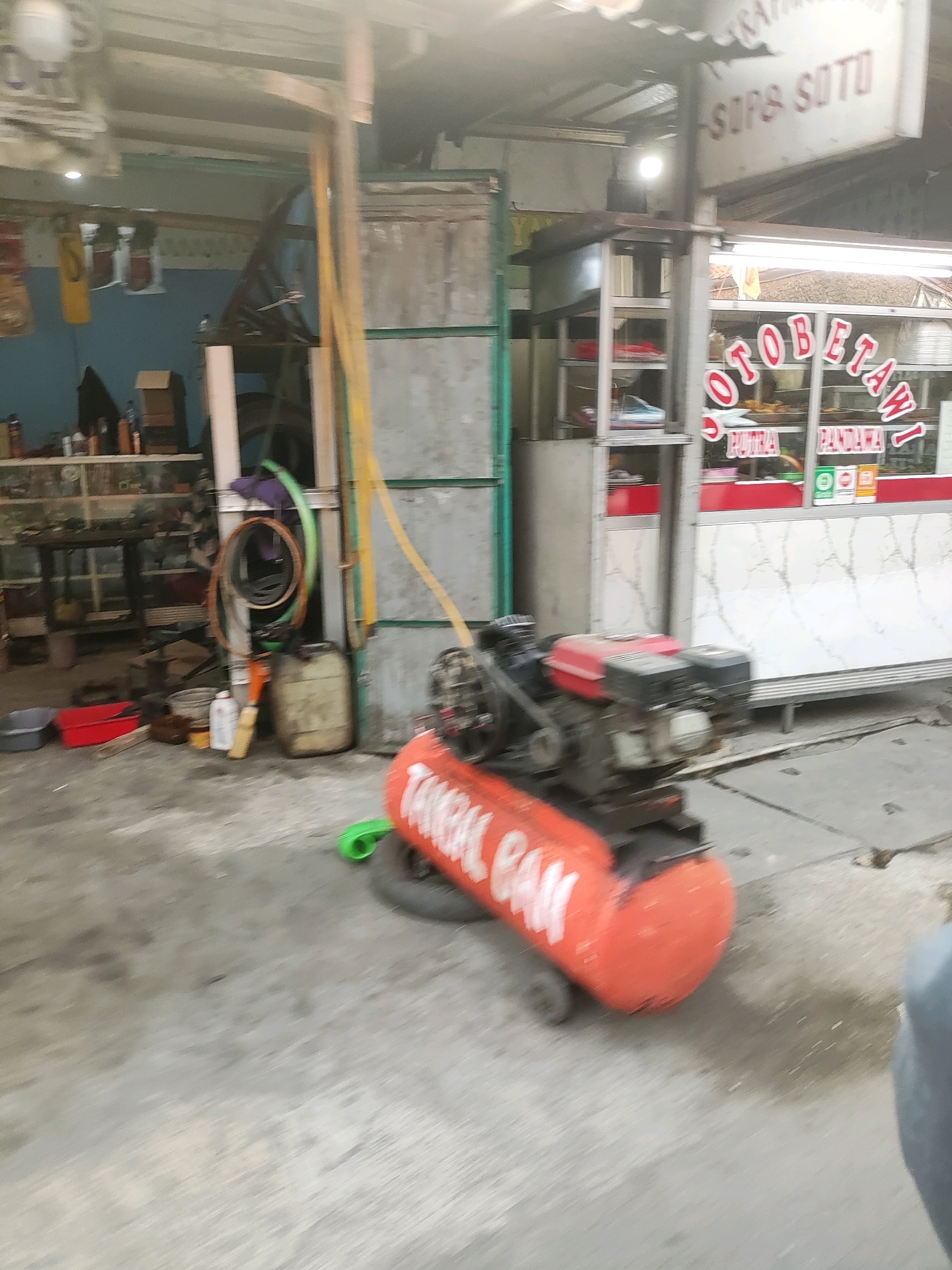 In Wanajaya, Indonesia, an outdoor scene depicts a collection of objects and structures suggesting a workspace or storage area. In the foreground, a red air compressor with white lettering is positioned on the ground, resting on small wheels. To its left, various containers and tools are scattered on a concrete floor. Behind the compressor, a yellow hose extends upwards and to the left. Further back, shelves stocked with items are visible against a blue wall. A metal structure with a corrugated roof is to the right, displaying signage that reads "SOTO SOPB." Adjacent to this structure is a shopfront with glass windows, featuring red trim and the prominent lettering "OTOBETAW," with "PUTRA PANDAWA" displayed below. The overall environment appears to be an open, somewhat cluttered outdoor area.